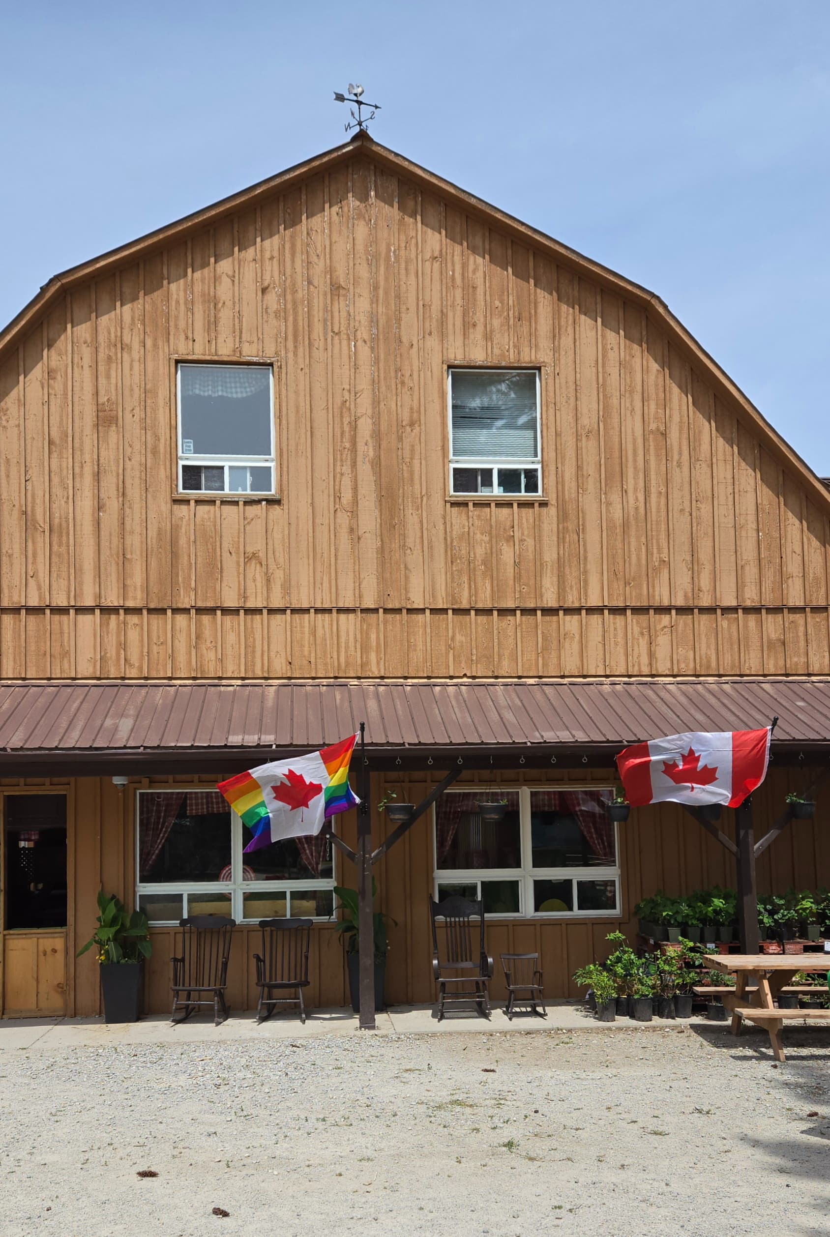 The front of the wooden farm market barn, with rocking chairs on the porch, potted plants and a Canadian flag and a Pride maple leaf flag