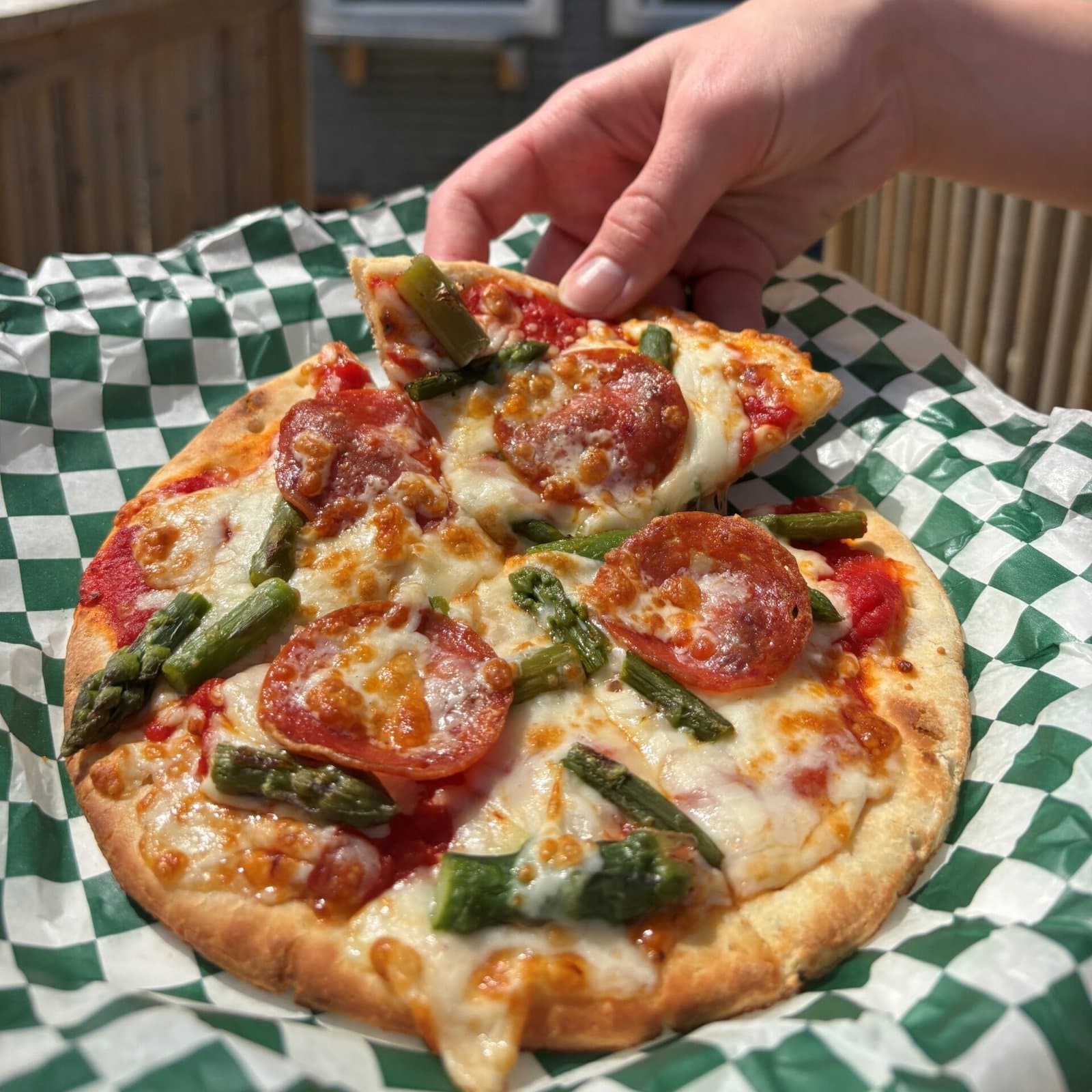 A hand lifts a slice from a pepperoni and asparagus pizza on green and white checked paper at the Pizza Pit