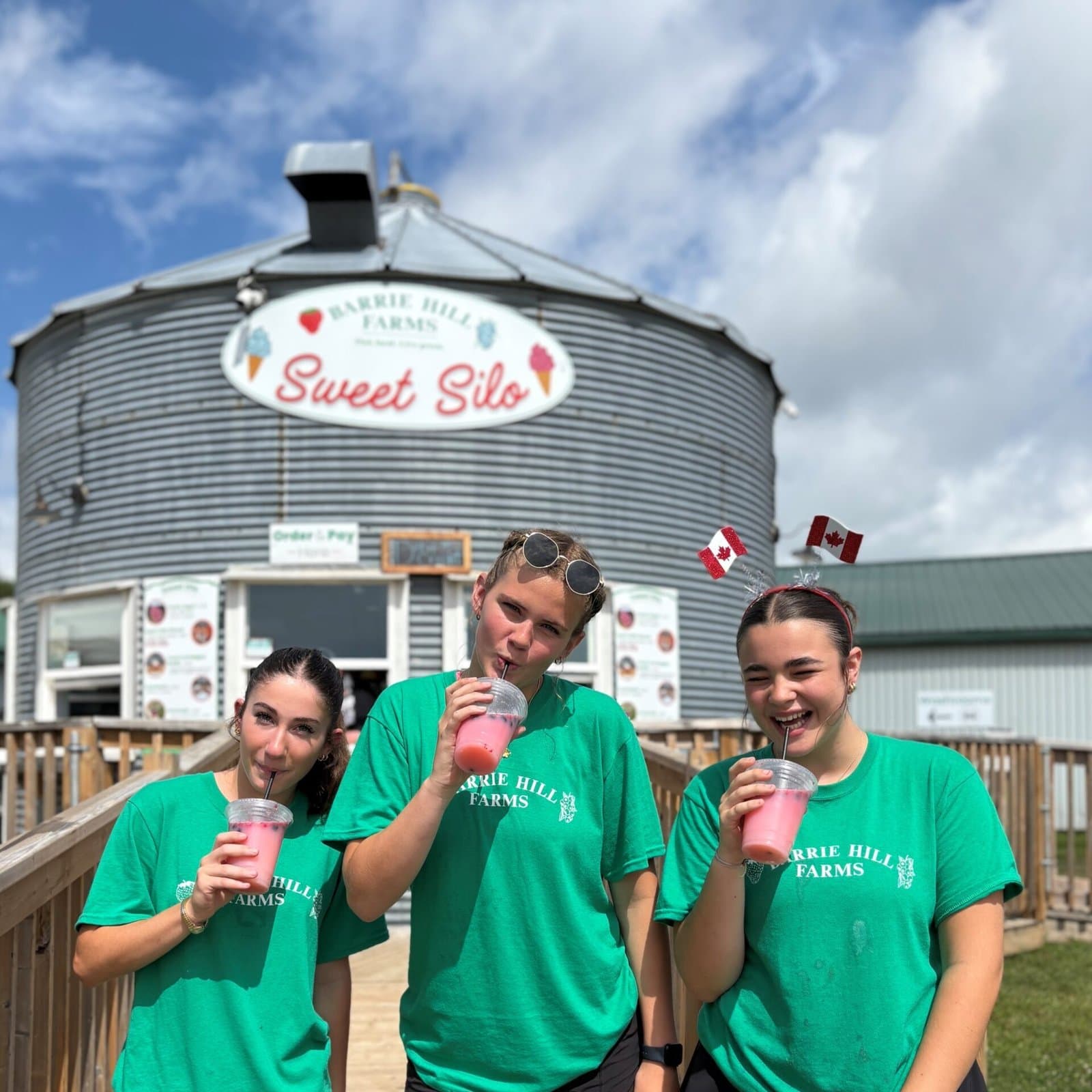 Three smiling staff in green Barrie Hill Farms T-shirts sip pink lemonade in front of the Sweet Silo, a grey grain bin turned snack counter