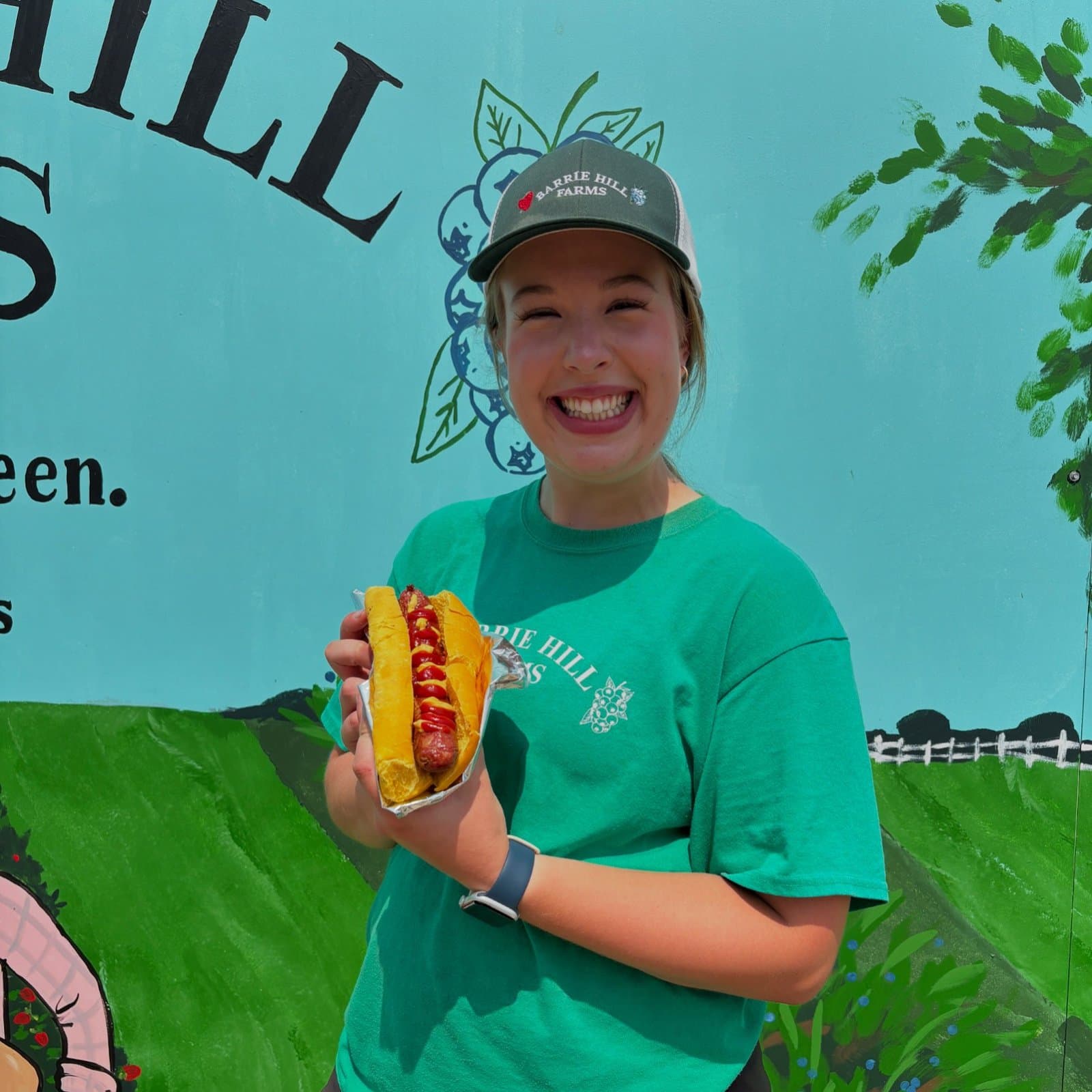 A grinning staff member in a Barrie Hill Farms cap and green T-shirt holds a grilled sausage on a bun in front of a painted farm mural