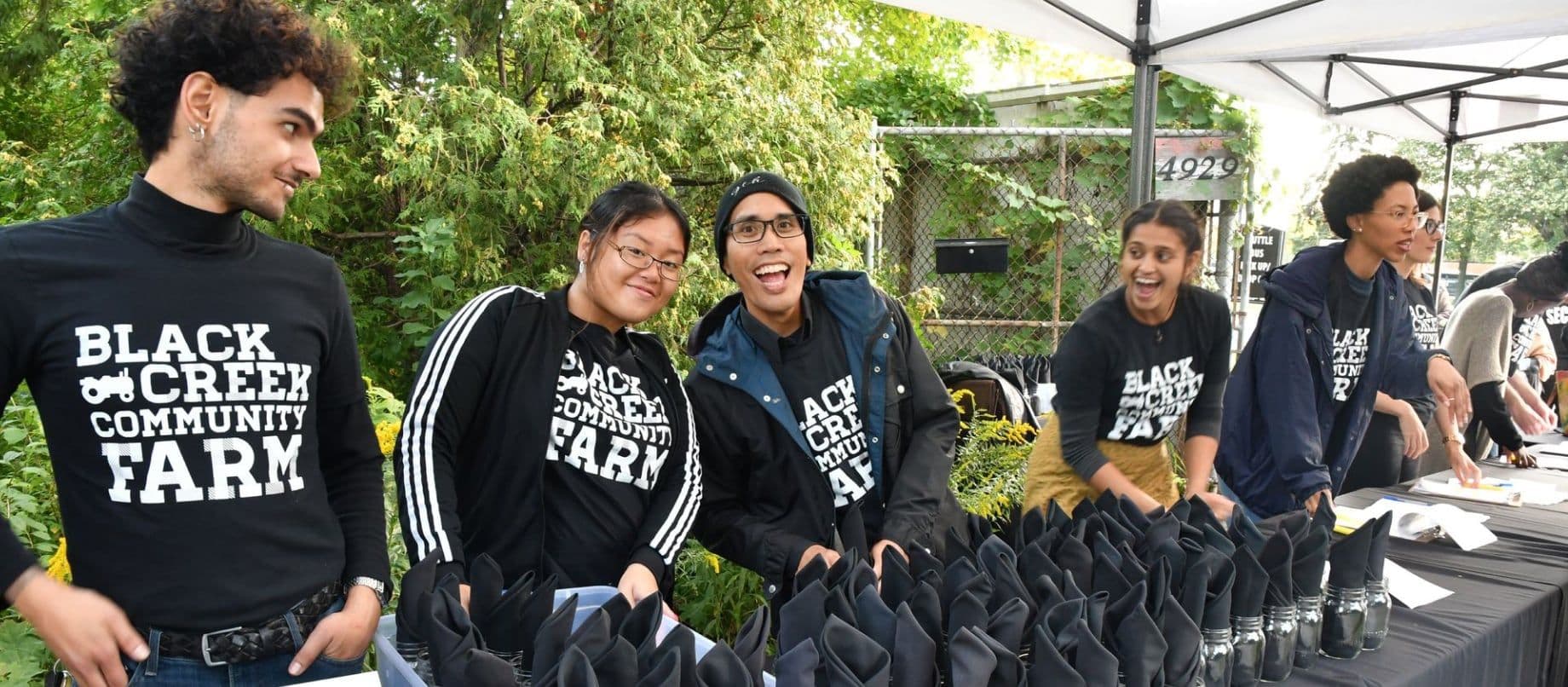 Smiling farm team members in Black Creek Community Farm shirts set out folded napkins on a table under a tent, with the 4929 gate sign behind