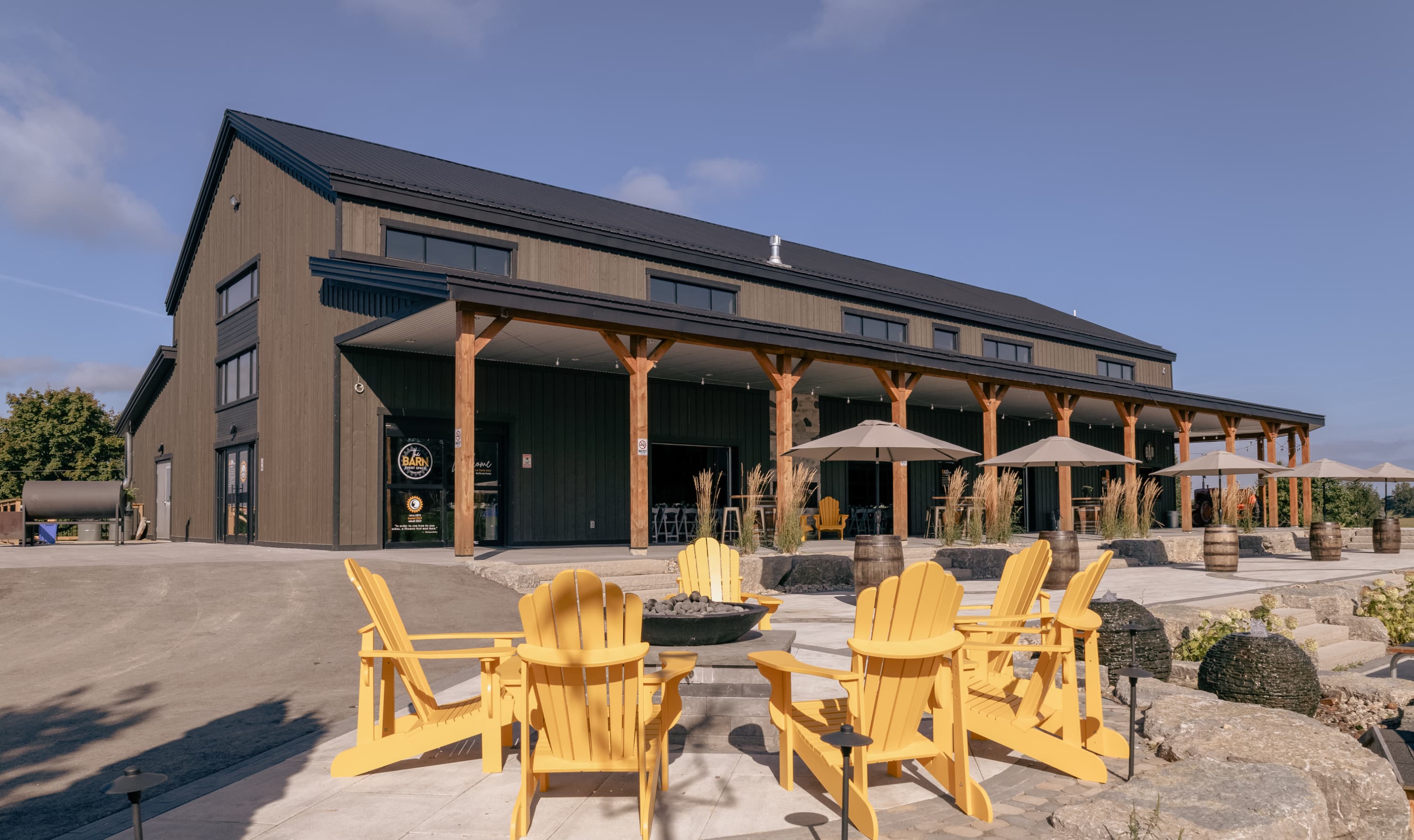The farm's rebuilt dark timber barn with a covered patio, umbrellas and yellow Muskoka chairs around a fire pit