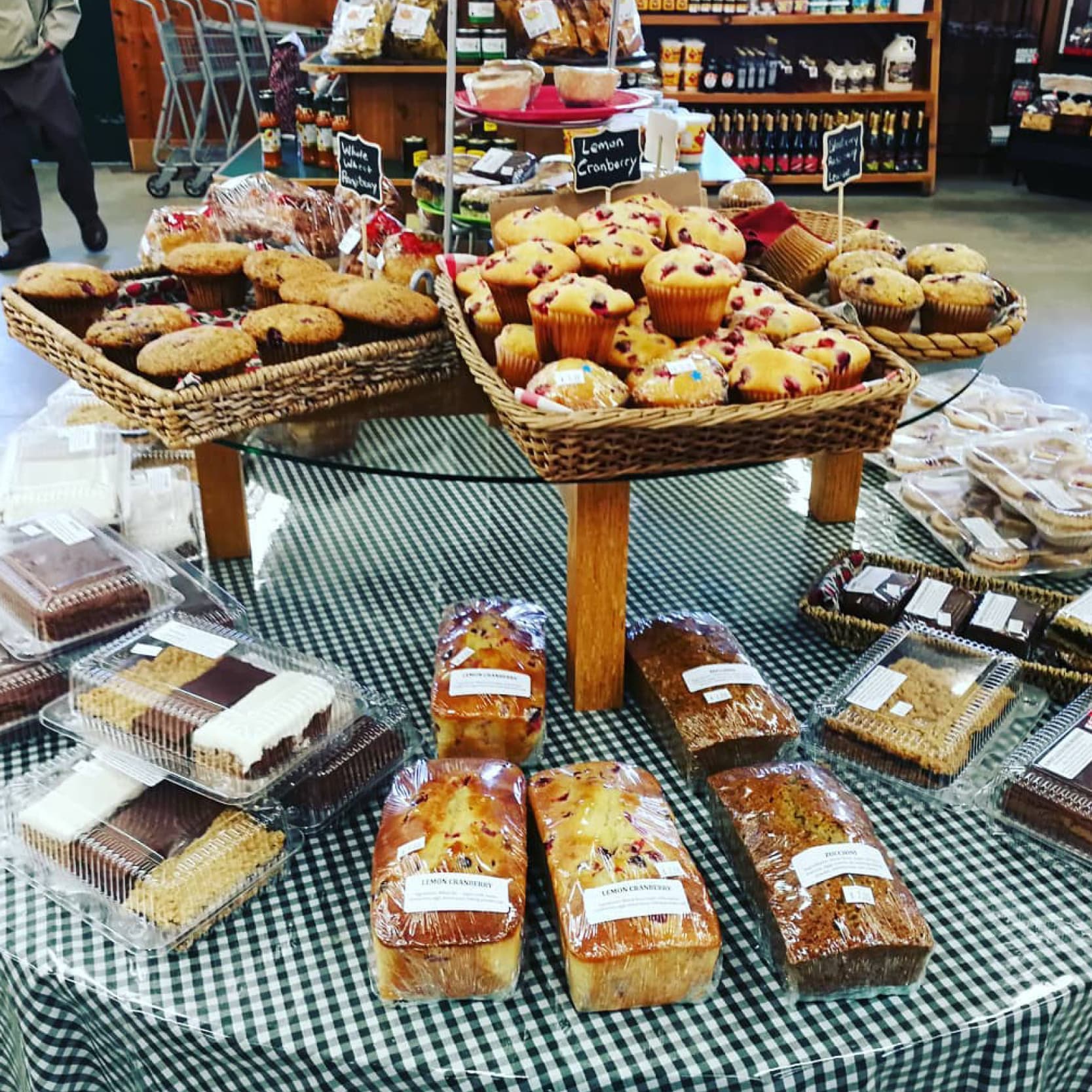 A gingham-covered table in the market laid with baskets of cranberry muffins, lemon cranberry and zucchini loaves and boxed squares, with chalkboard labels