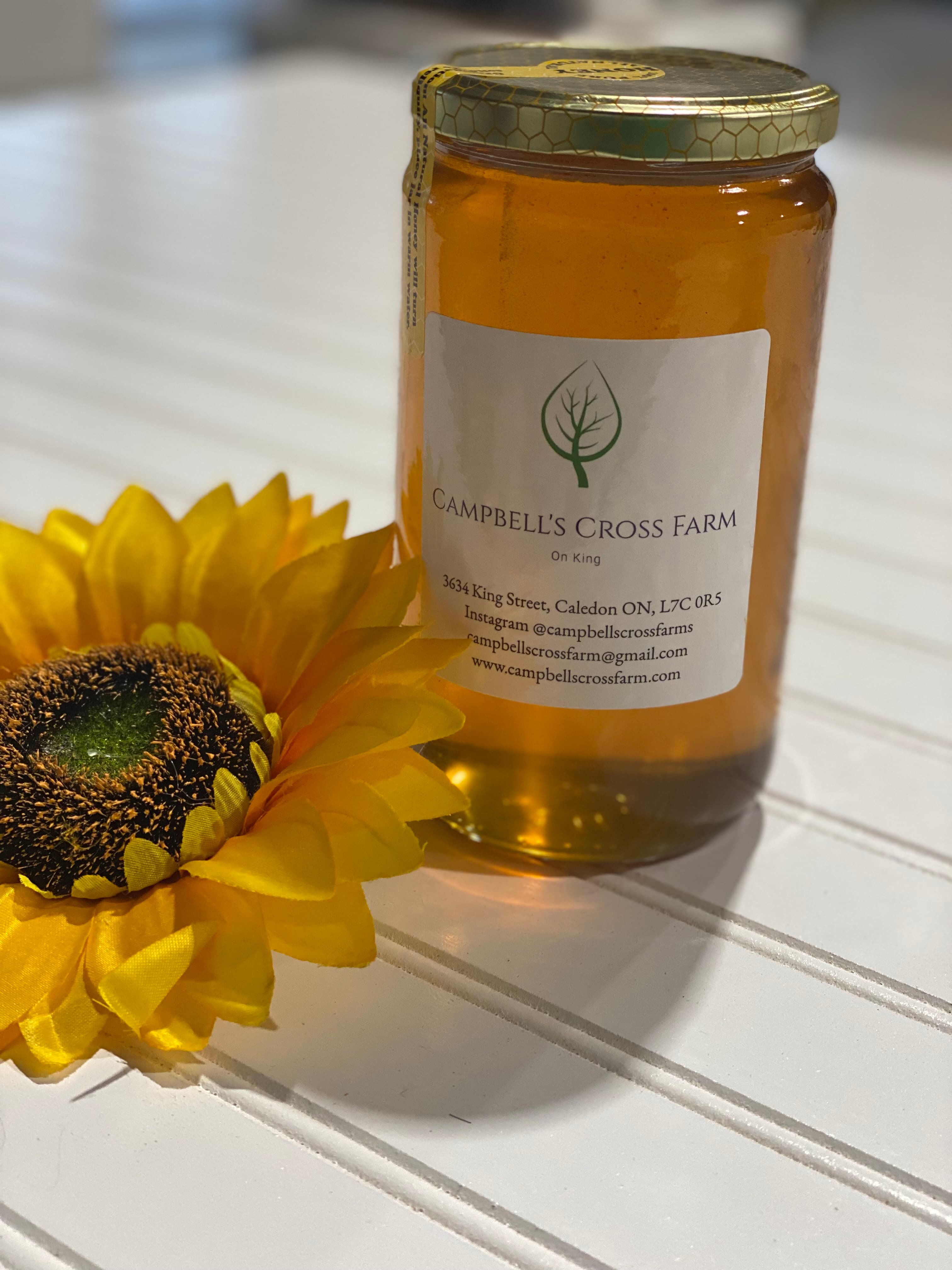 A glass jar of golden Campbell's Cross Farm honey with the farm's leaf label, beside a sunflower bloom on a white table