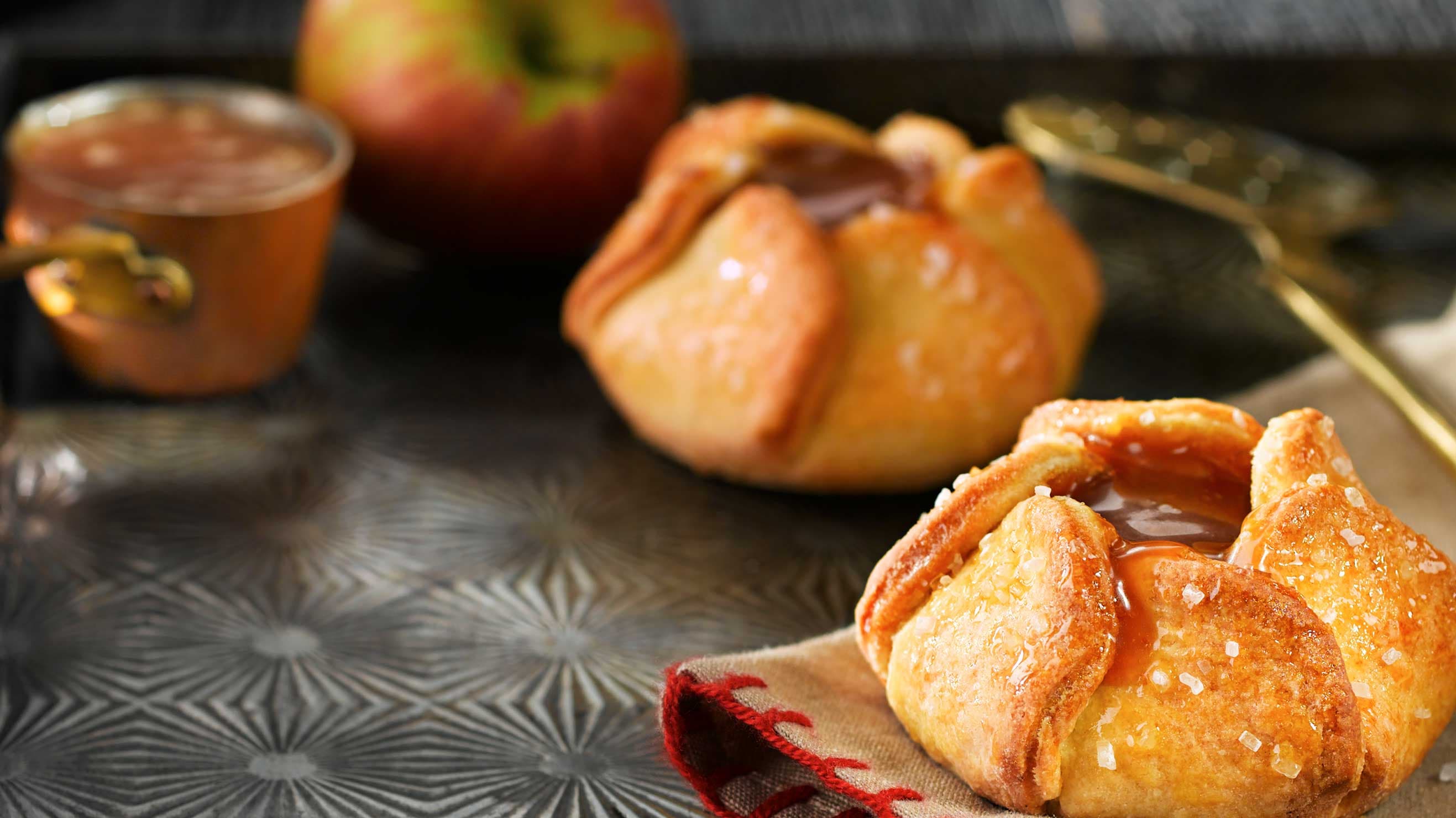 Two baked apple blossom pastries topped with caramel and coarse sugar, with an apple and a pot of caramel behind