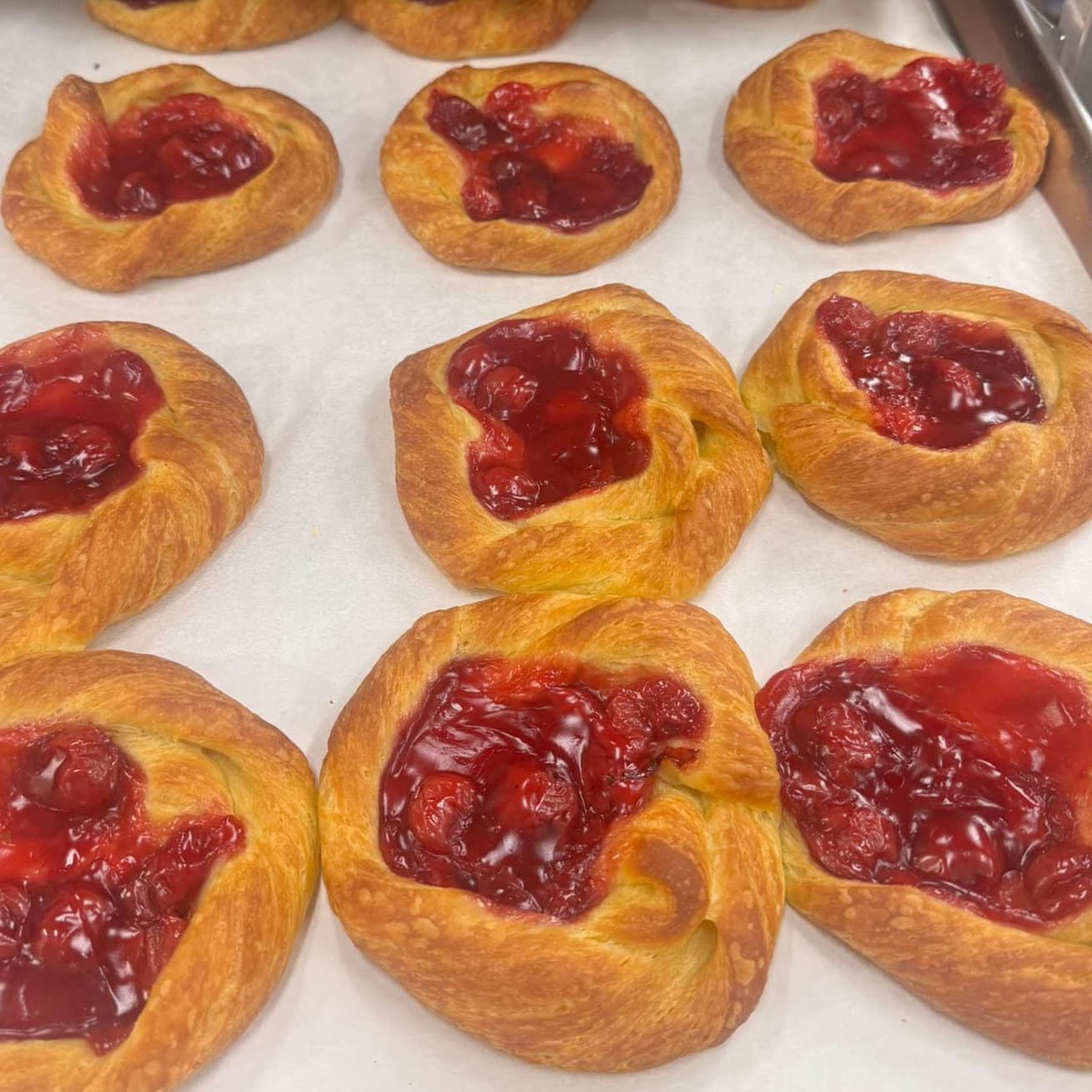 A baking tray of golden danishes filled with cherry, cooling on parchment