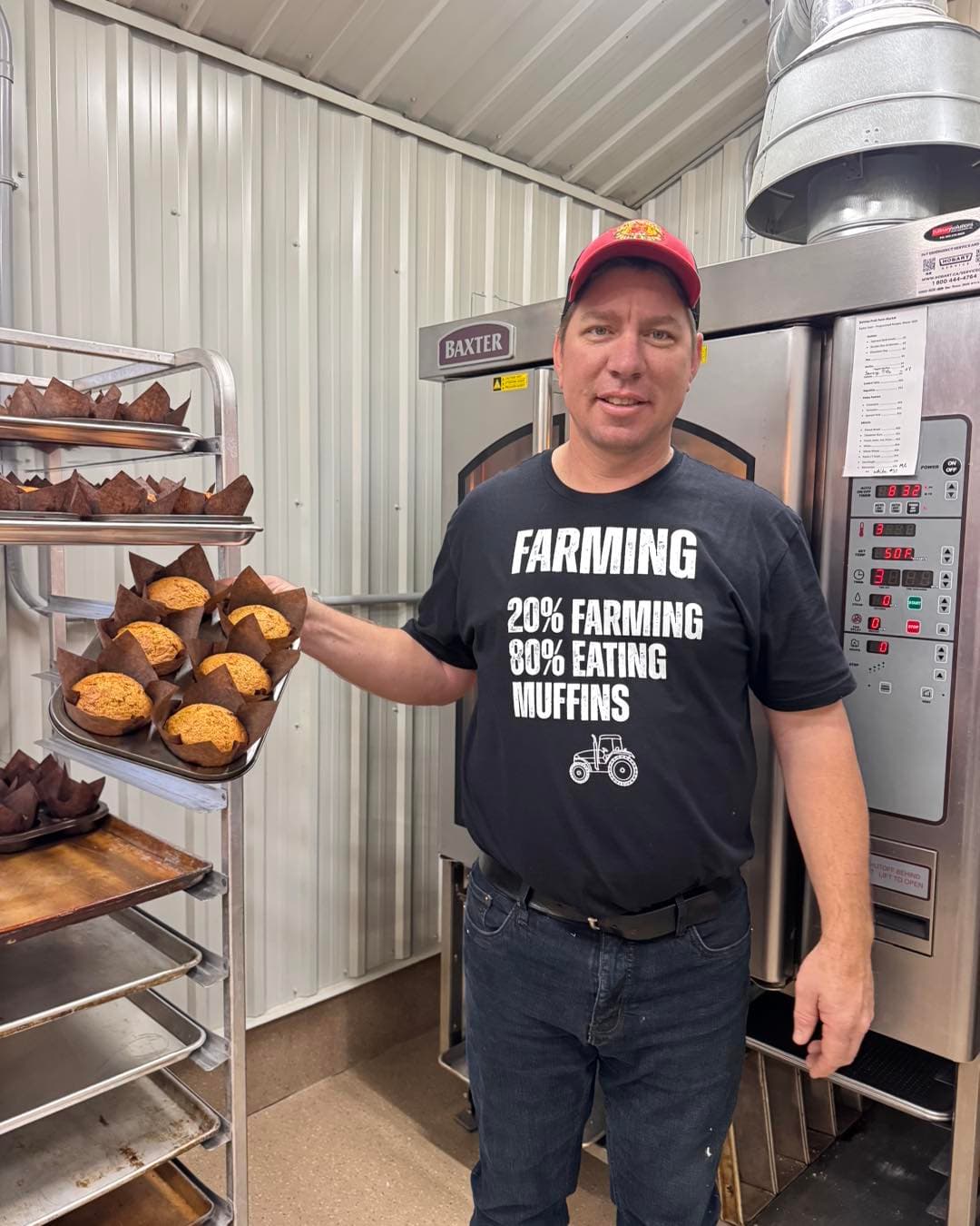 A smiling man in a red cap holds out a tray of fresh muffins beside the bakery's ovens