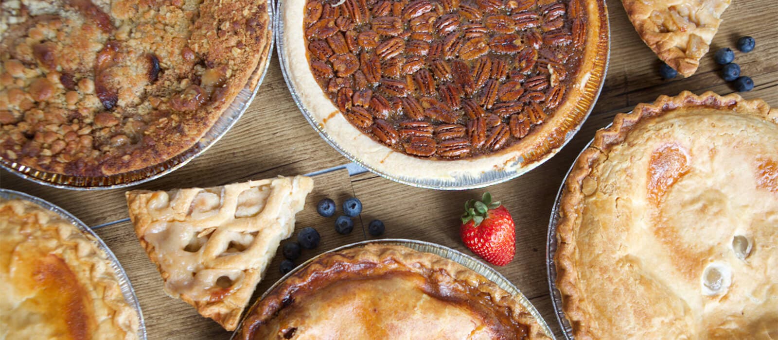 Pecan, crumble, lattice and double-crust pies in foil tins on a wooden table, with blueberries and a strawberry between them