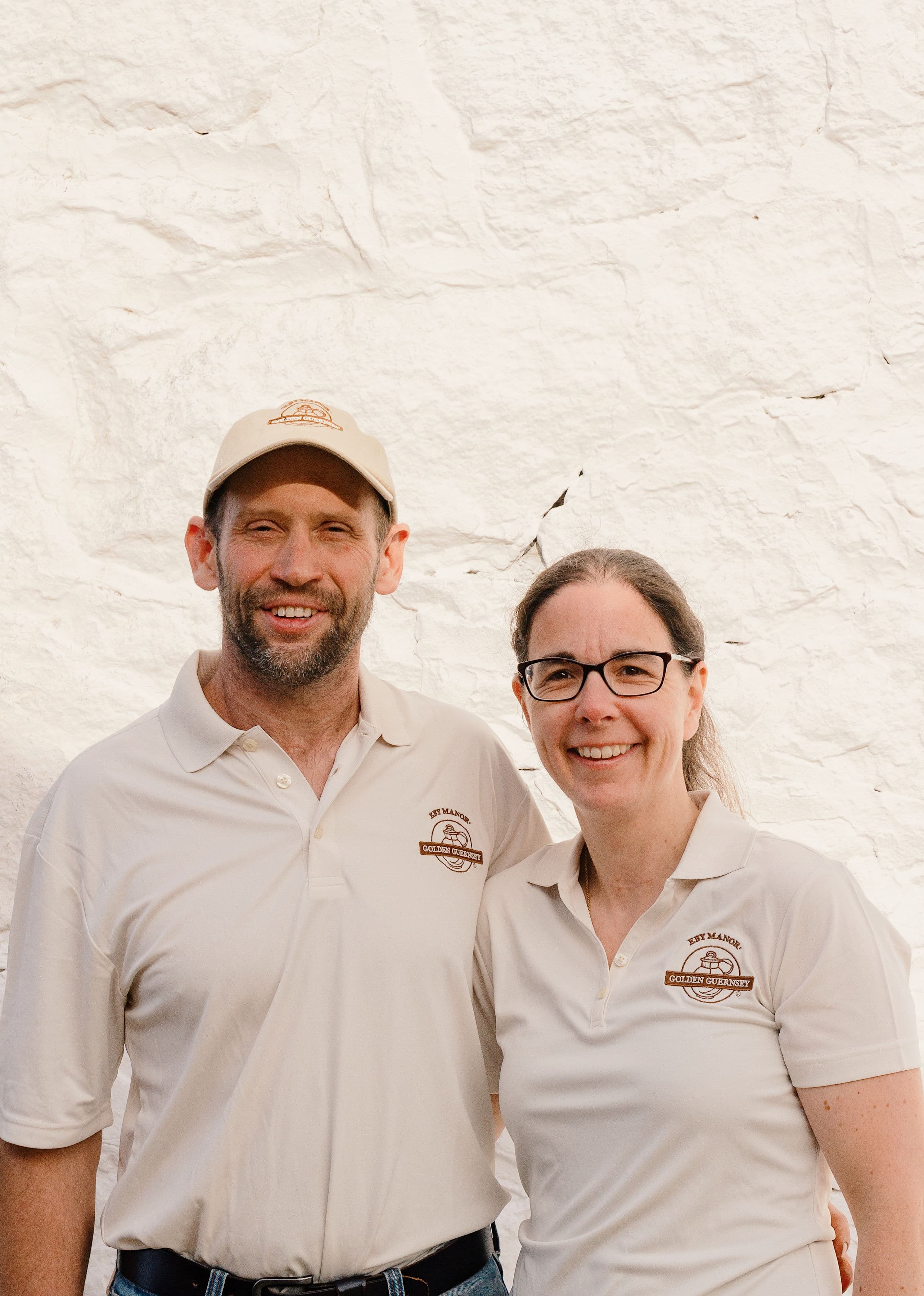 Co-owners Ben and Sheri Eby smiling in Eby Manor Golden Guernsey shirts against a whitewashed stone wall