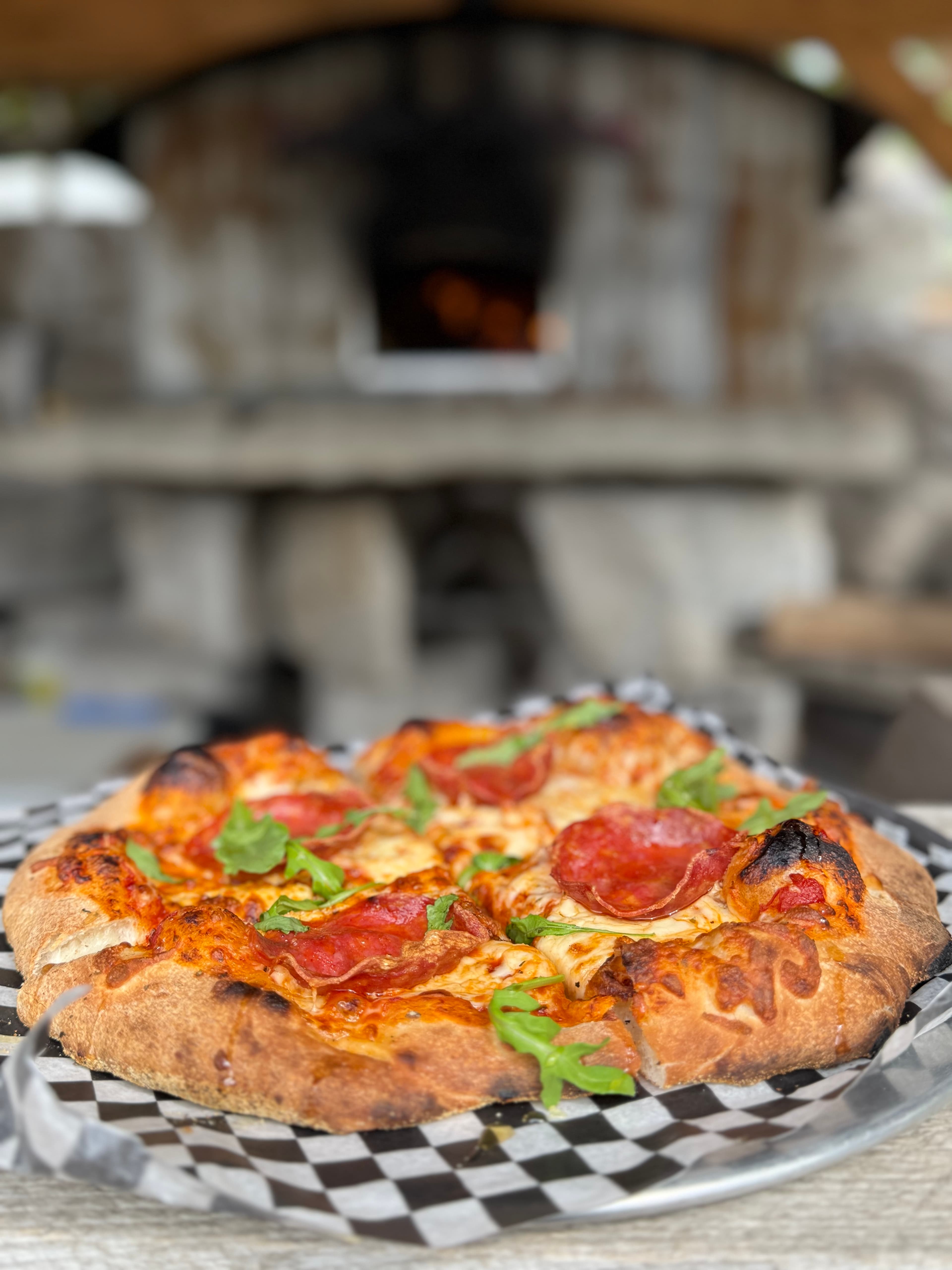 A wood-fired pizza topped with salami and fresh arugula on a checkered paper liner, with the cafe's stone oven glowing behind it