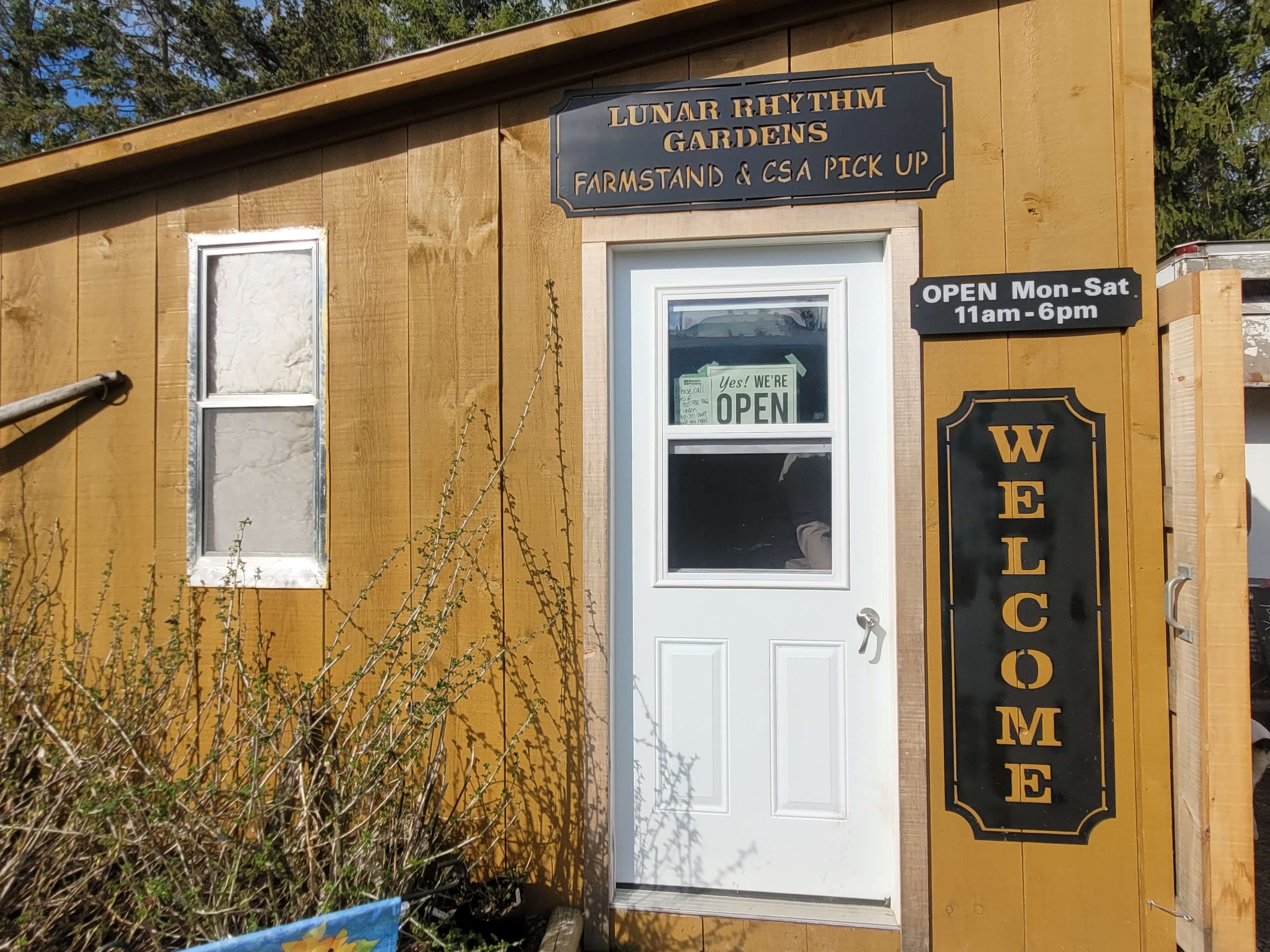 The mustard-yellow board-and-batten farm stand, with a white door under a black sign reading Lunar Rhythm Gardens Farmstand & CSA Pick Up, an OPEN Mon-Sat 11am-6pm sign and a tall WELCOME sign