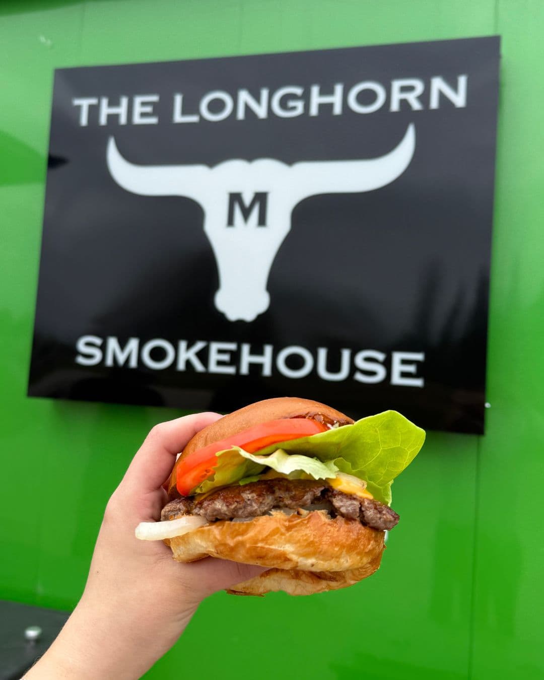A smash burger with lettuce and tomato held up in front of the Longhorn Smokehouse food truck sign