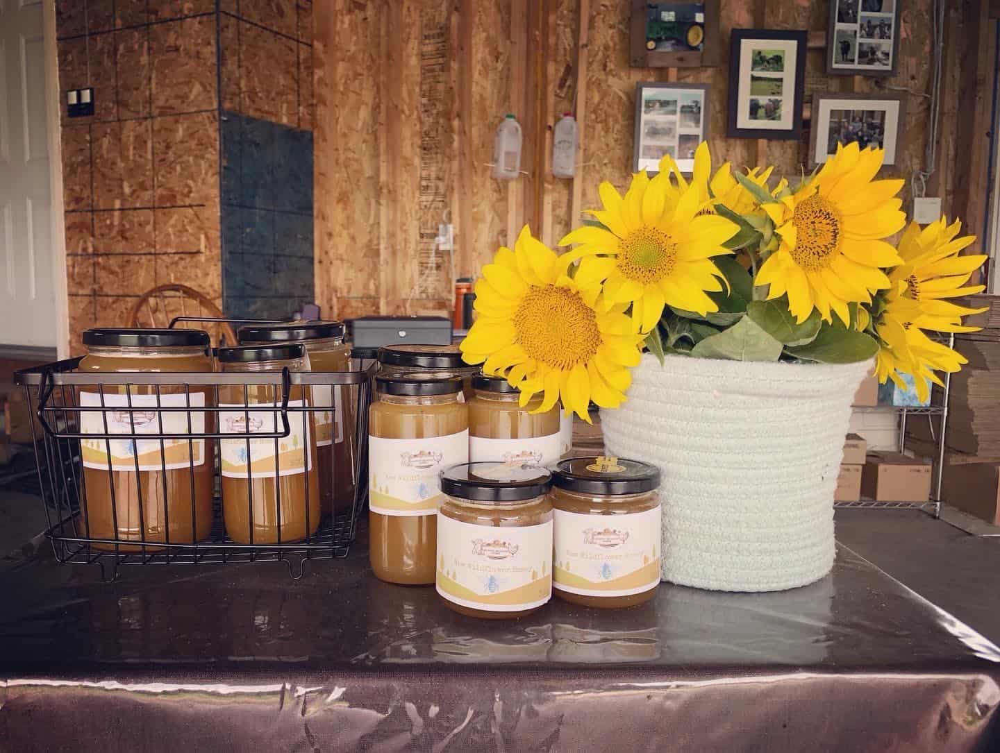 Jars of Murphy Meadow Farms raw wildflower honey beside a basket of sunflowers on a table against a wooden wall