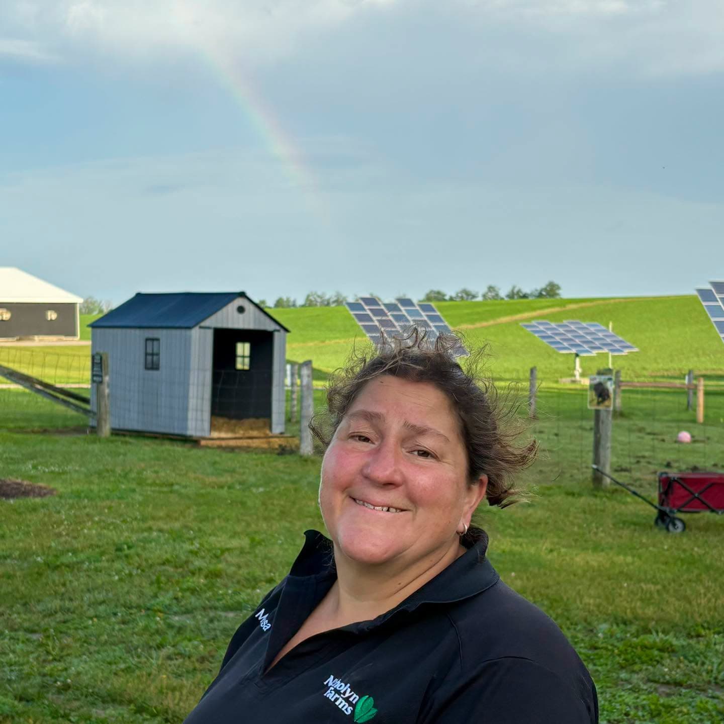 Melissa Van Casteren smiling in a Nicholyn Farms shirt by the animal paddock, with solar panels and a faint rainbow behind