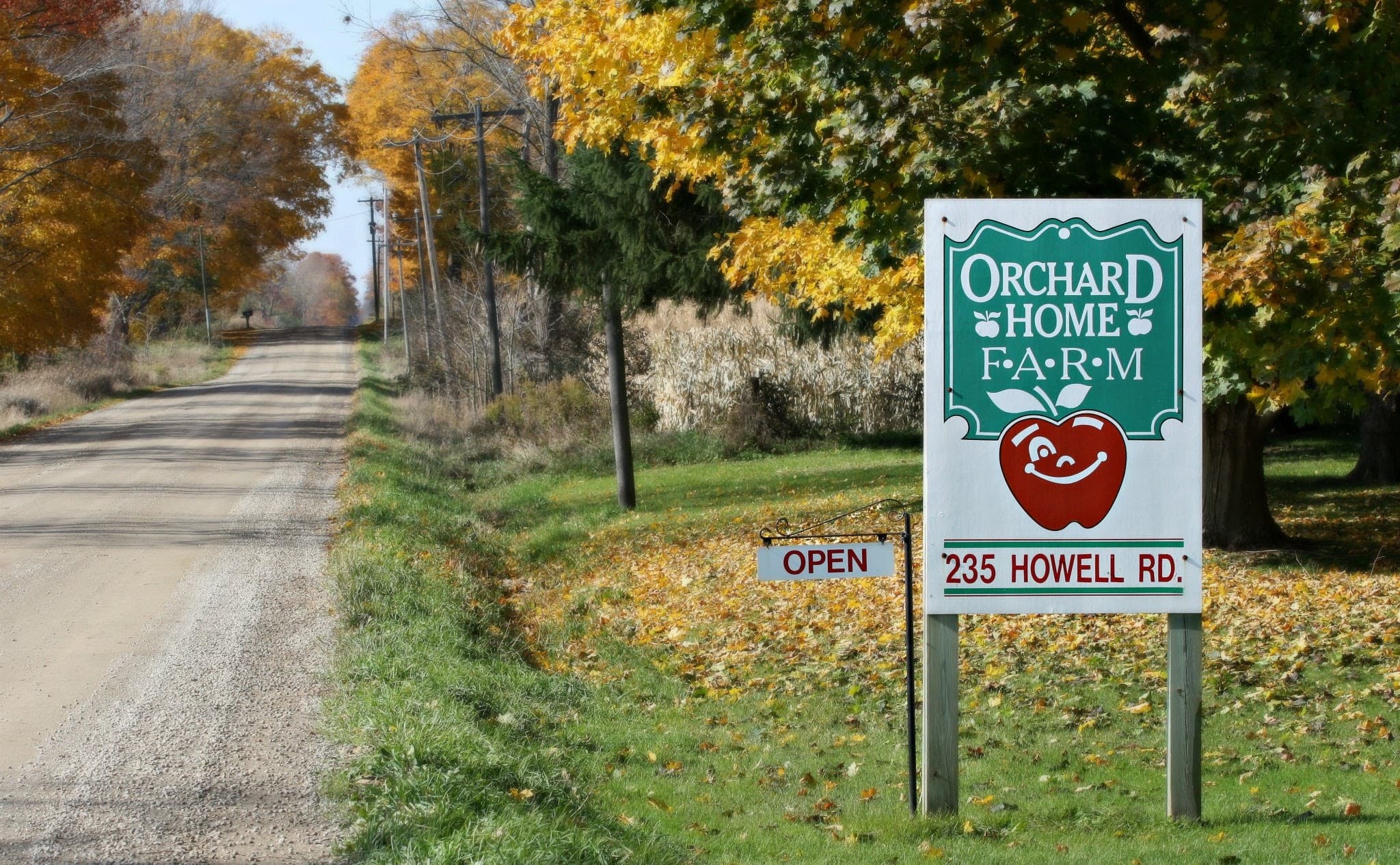 The green and white Orchard Home Farm sign, with a smiling red apple, an OPEN plate and 235 Howell Rd., beside the gravel road under autumn maples