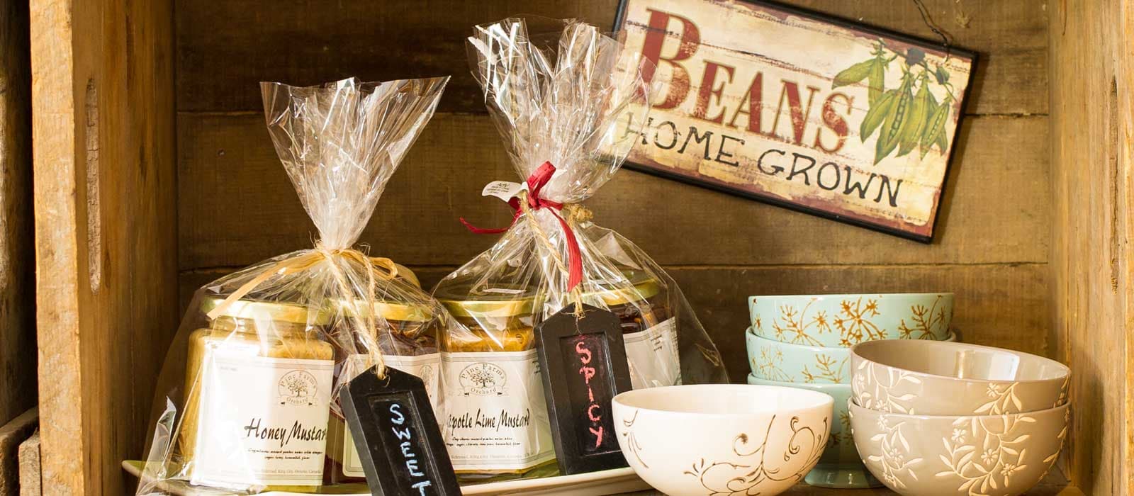 Jars of Pine Farms Orchard honey mustard and chipotle lime mustard wrapped with ribbon and chalkboard tags, beside stacked ceramic bowls on a wooden shelf