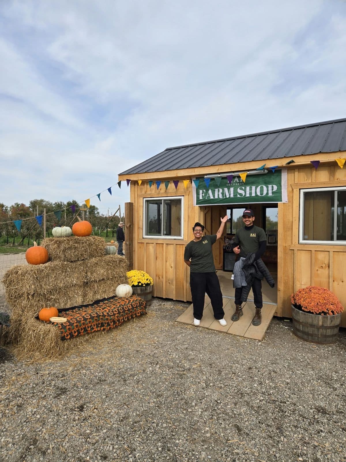 Two smiling team members in Rai Orchards T-shirts wave from the door of the wooden farm shop, beside hay bales stacked with pumpkins and tubs of mums