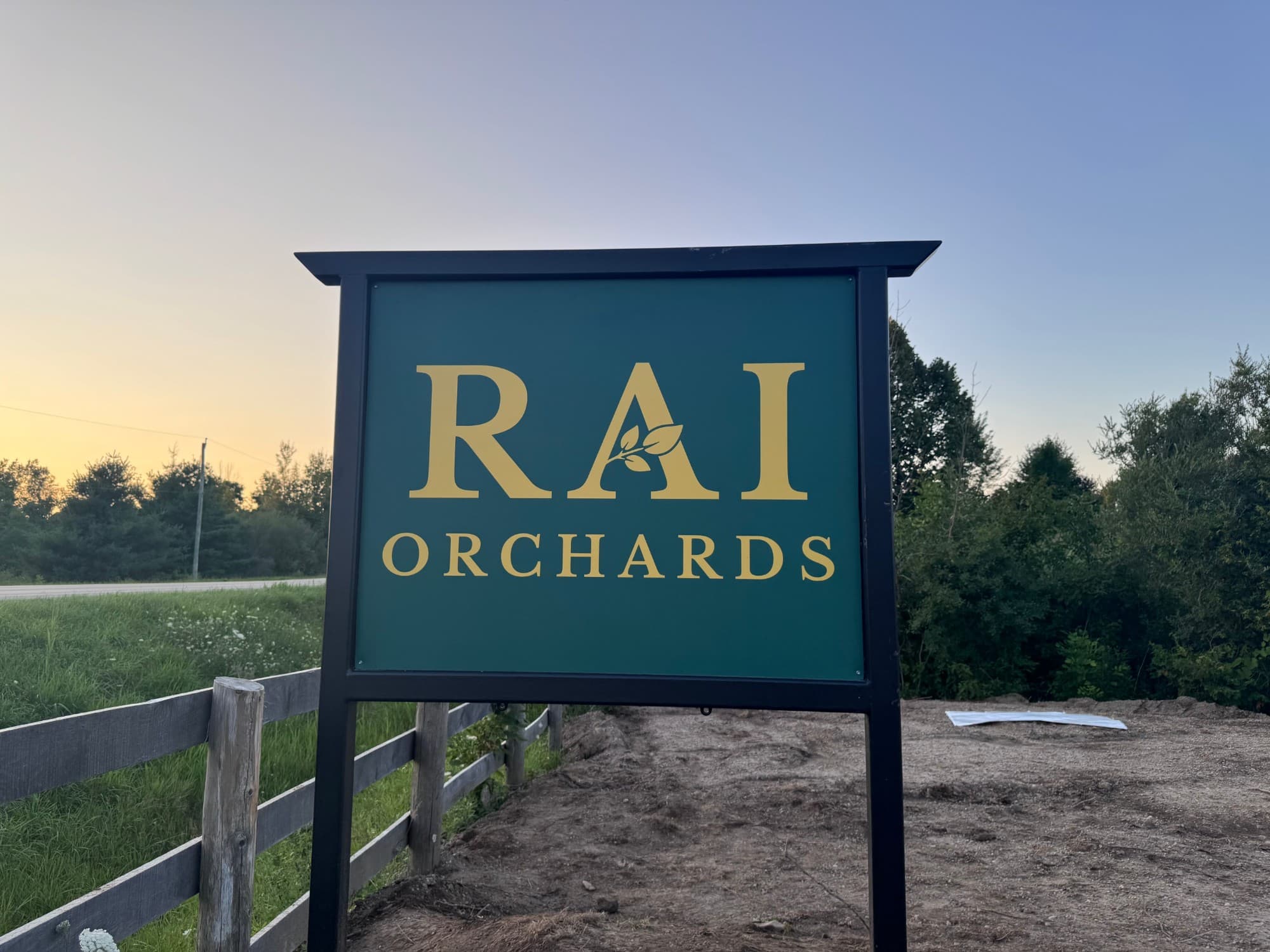 The green and gold Rai Orchards sign by a split-rail fence at the roadside, with trees and a sunset sky behind
