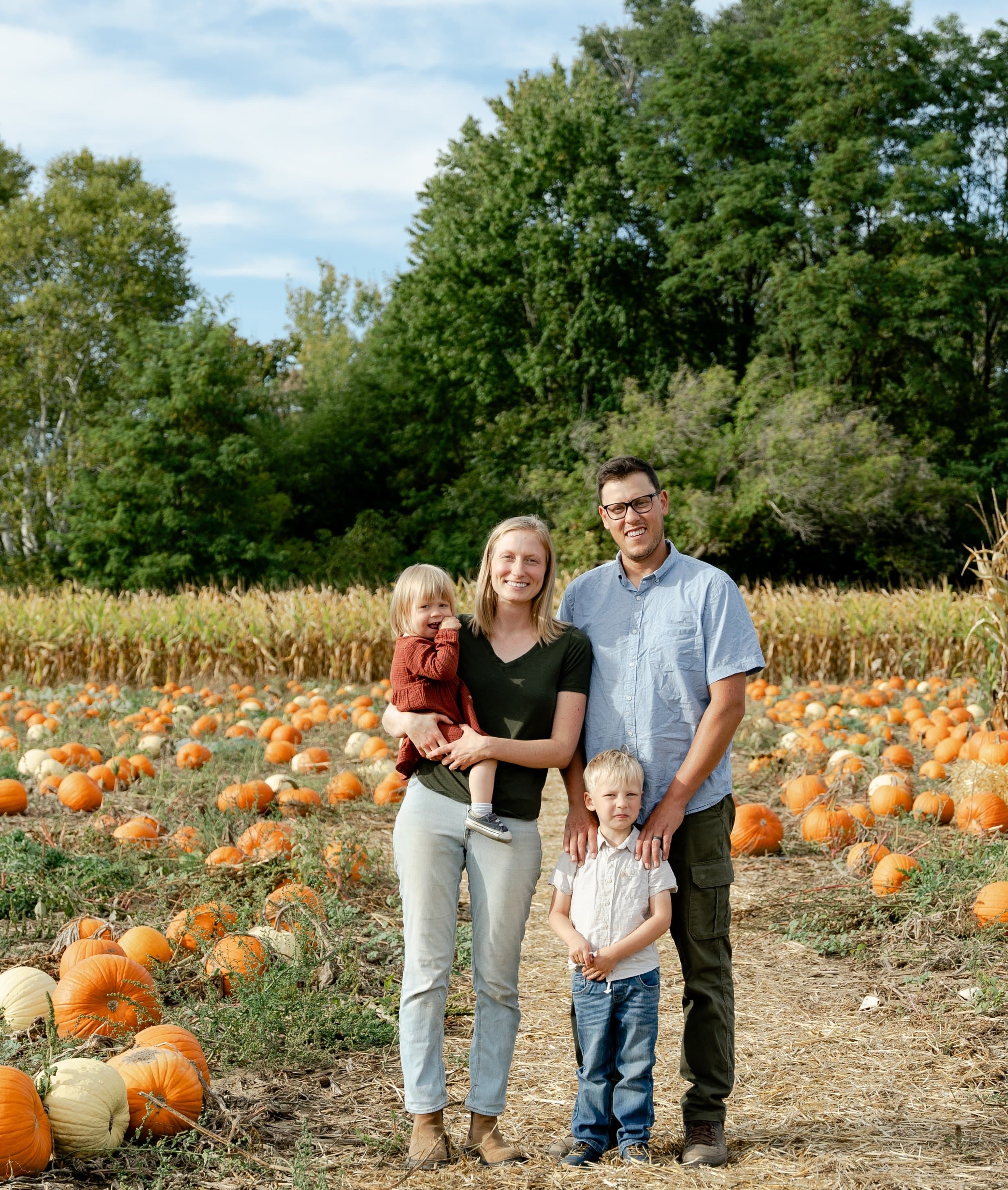 John and Emma Reesor smile with their two young children on a straw path through the pumpkin patch, with corn and trees behind