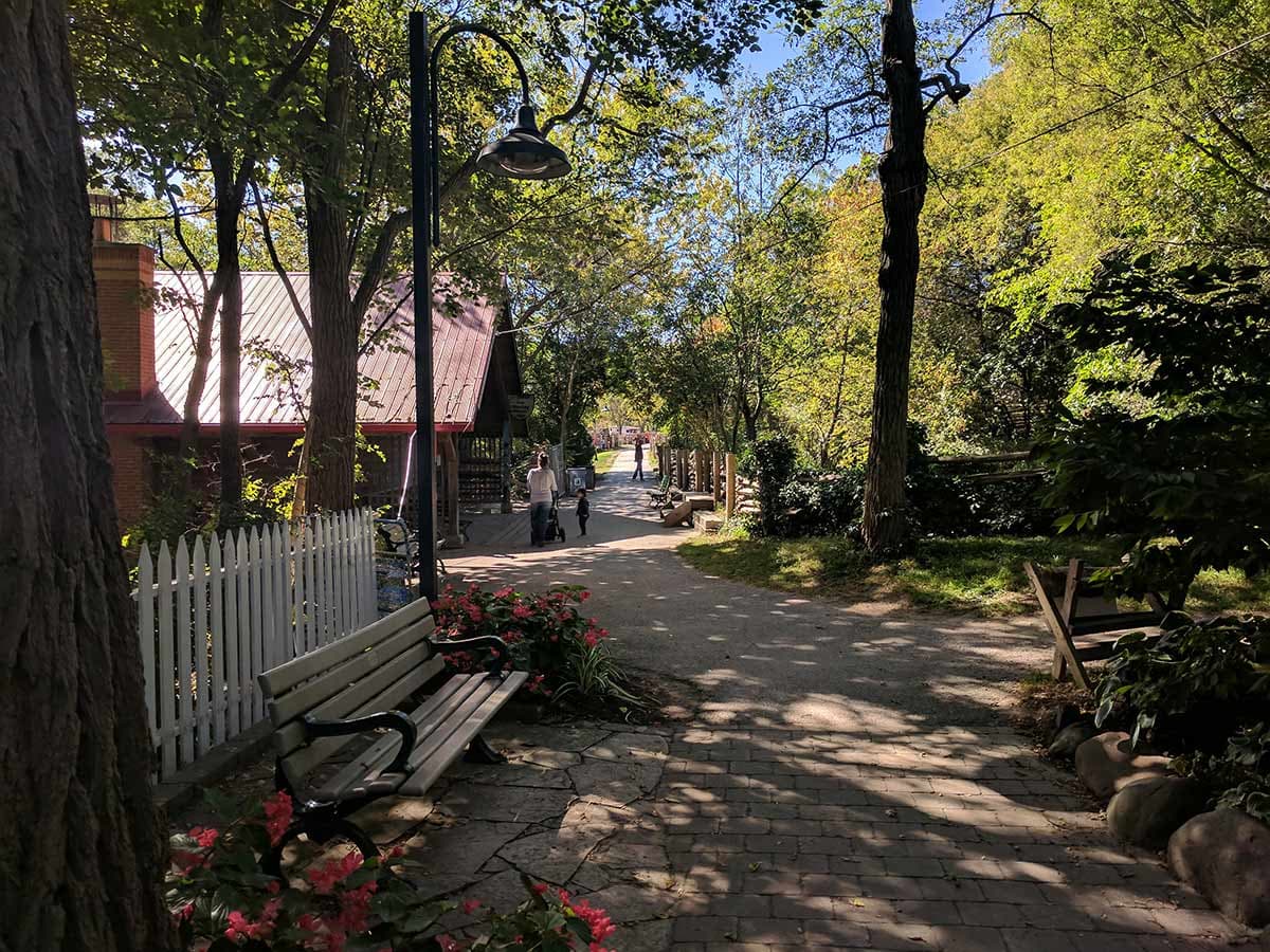 A brick building with a red metal roof and a white picket fence along a shaded paved lane with a bench and pink flowers