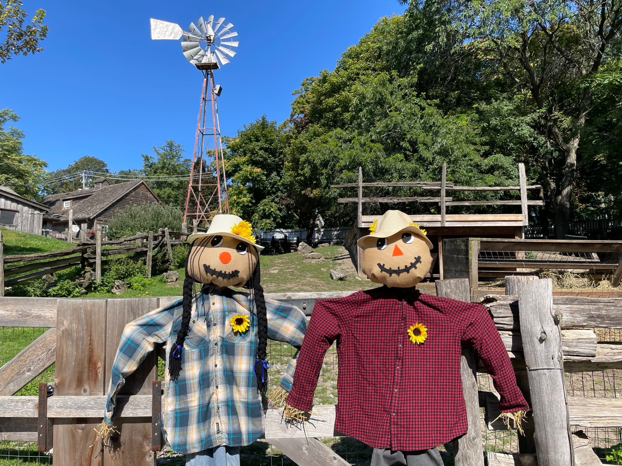 Two scarecrows in plaid shirts and straw hats lean on a split-rail fence, with a windmill, a weathered barn and the paddocks behind
