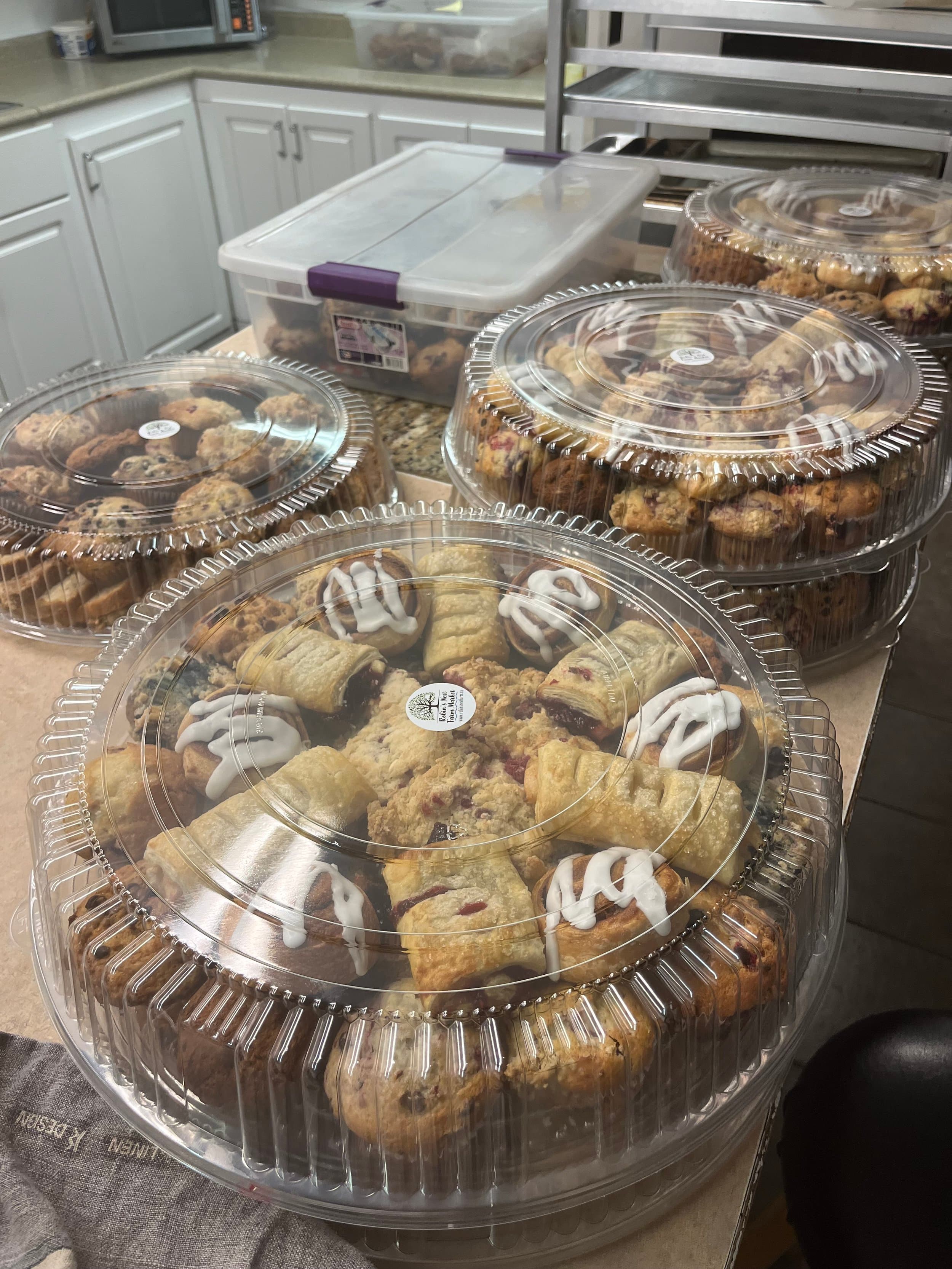 Covered trays of cinnamon buns, fruit turnovers, crumble squares and muffins with Robin's Nest Farm Market stickers, on the kitchen counter