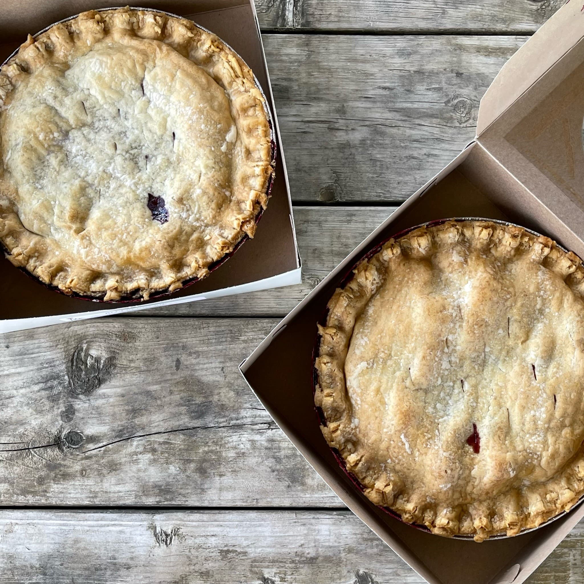 Two golden double-crust fruit pies in open bakery boxes on a weathered wooden table