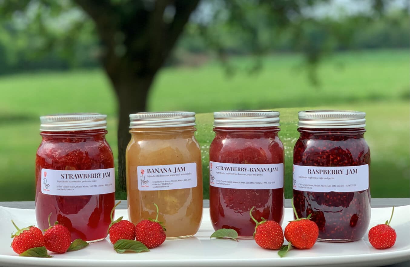 Four jars of the farm's strawberry, banana, strawberry-banana and raspberry jam with fresh strawberries, outdoors under a tree