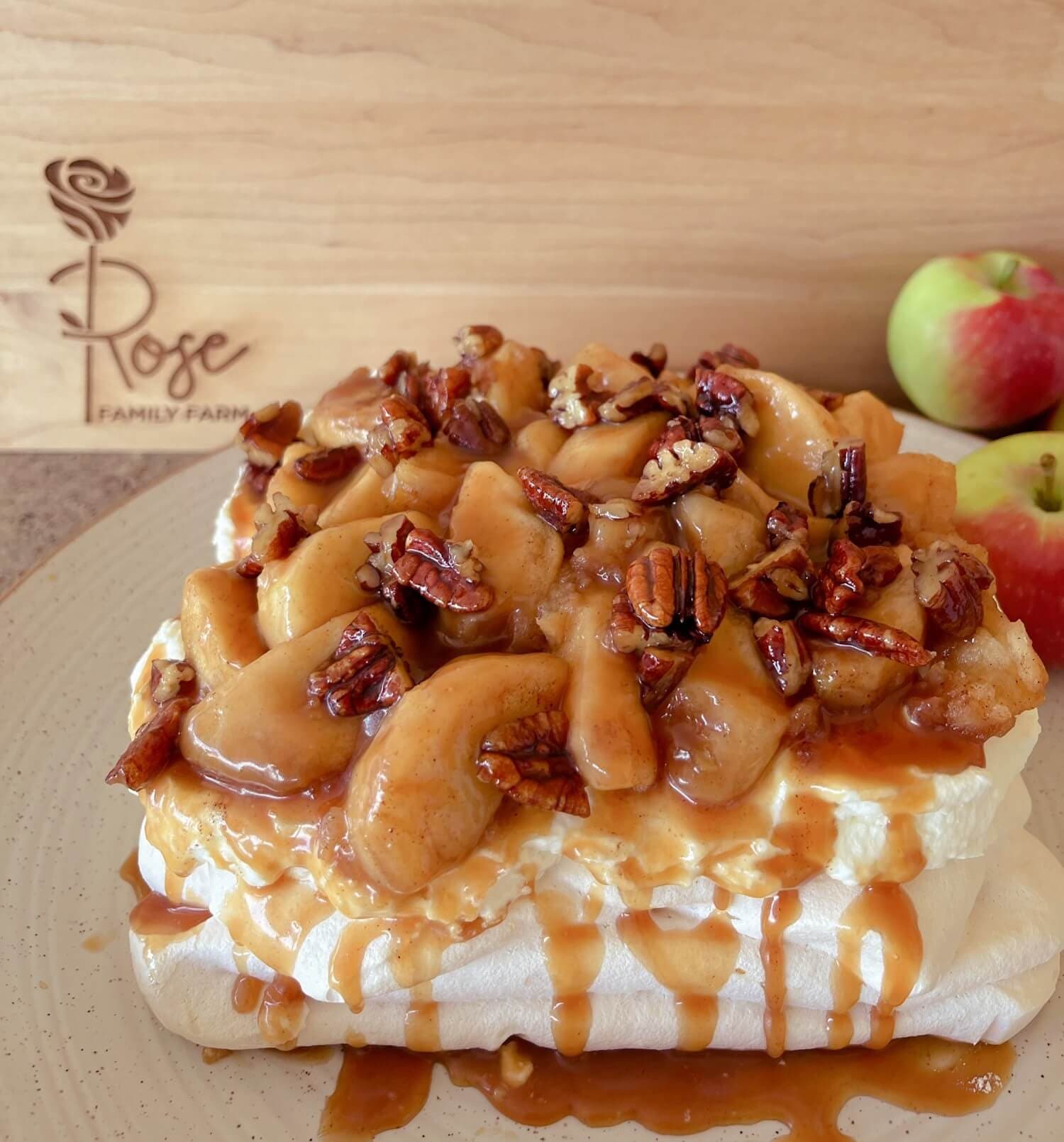 An apple caramel pecan pavlova dripping with caramel, in front of a wooden board engraved with the Rose Family Farm logo