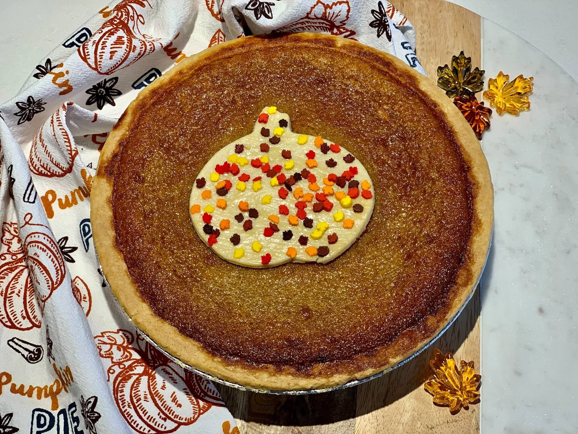 A whole pumpkin pie topped with a pastry pumpkin and leaf sprinkles, on a wooden board beside a pumpkin-print tea towel
