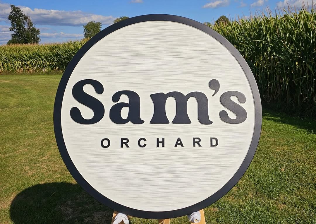 The round white Sam's Orchard sign stands on the lawn in front of a tall field of corn under a blue sky