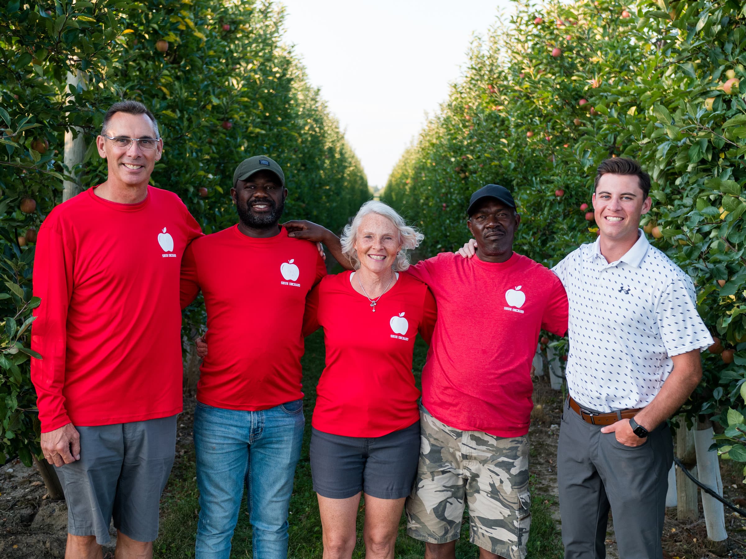 Five smiling members of the Shuh Orchards family and crew, four in red Shuh Orchards shirts, arm in arm between rows of apple trees heavy with red apples