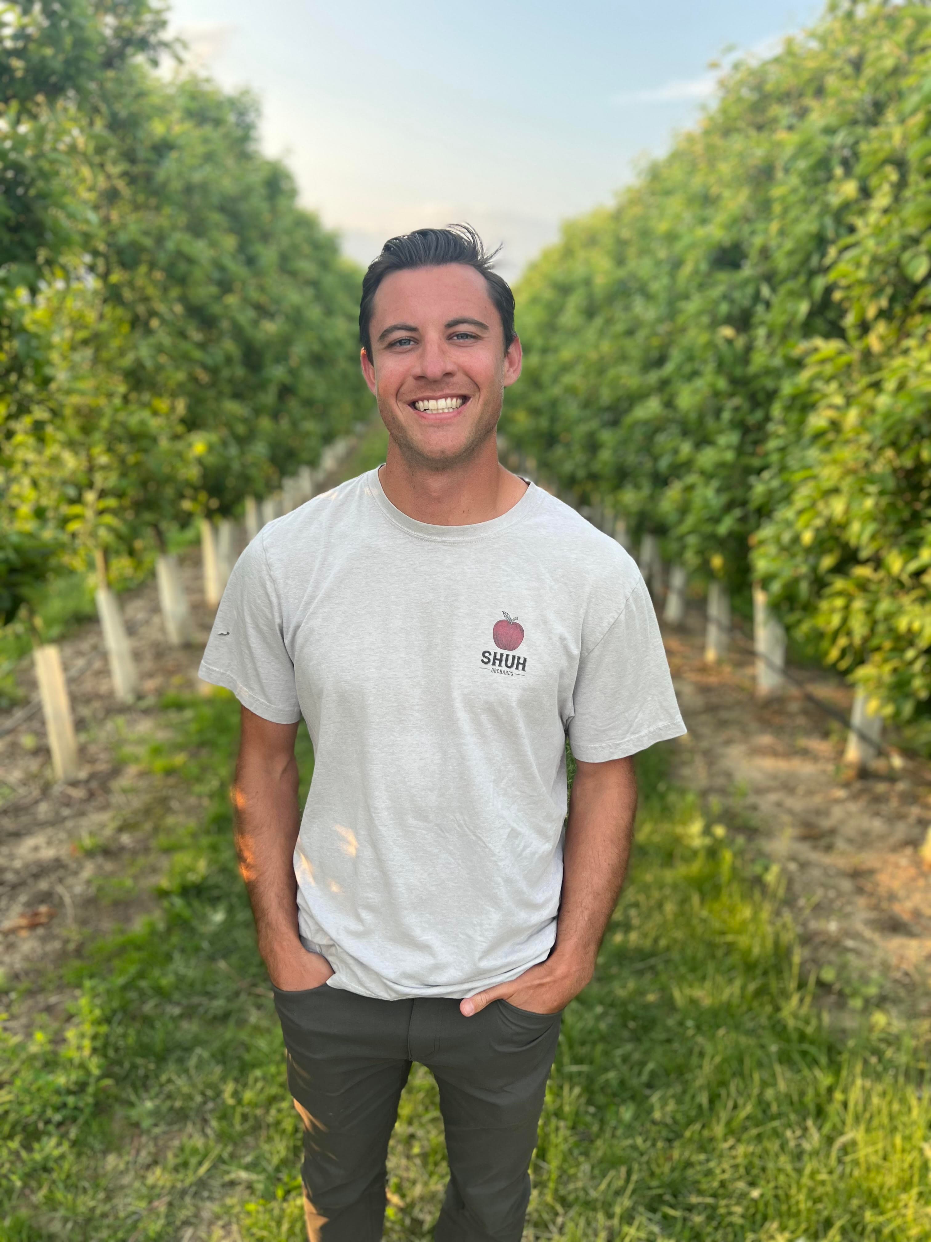 Tim Shuh, smiling in a Shuh Orchards T-shirt, stands between rows of young trellised apple trees