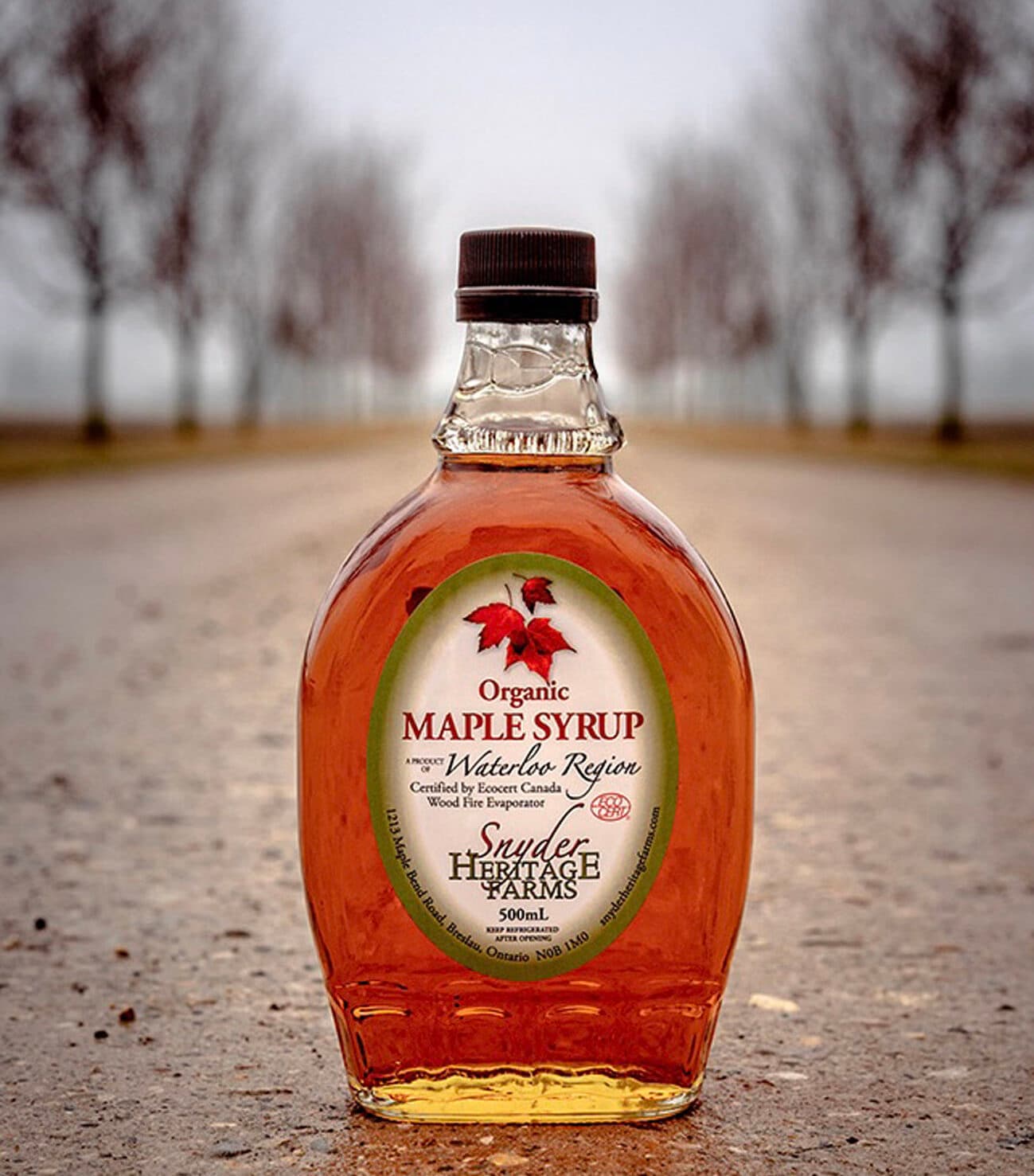 A 500 ml glass bottle of the farm's maple syrup standing on a gravel lane lined with bare trees