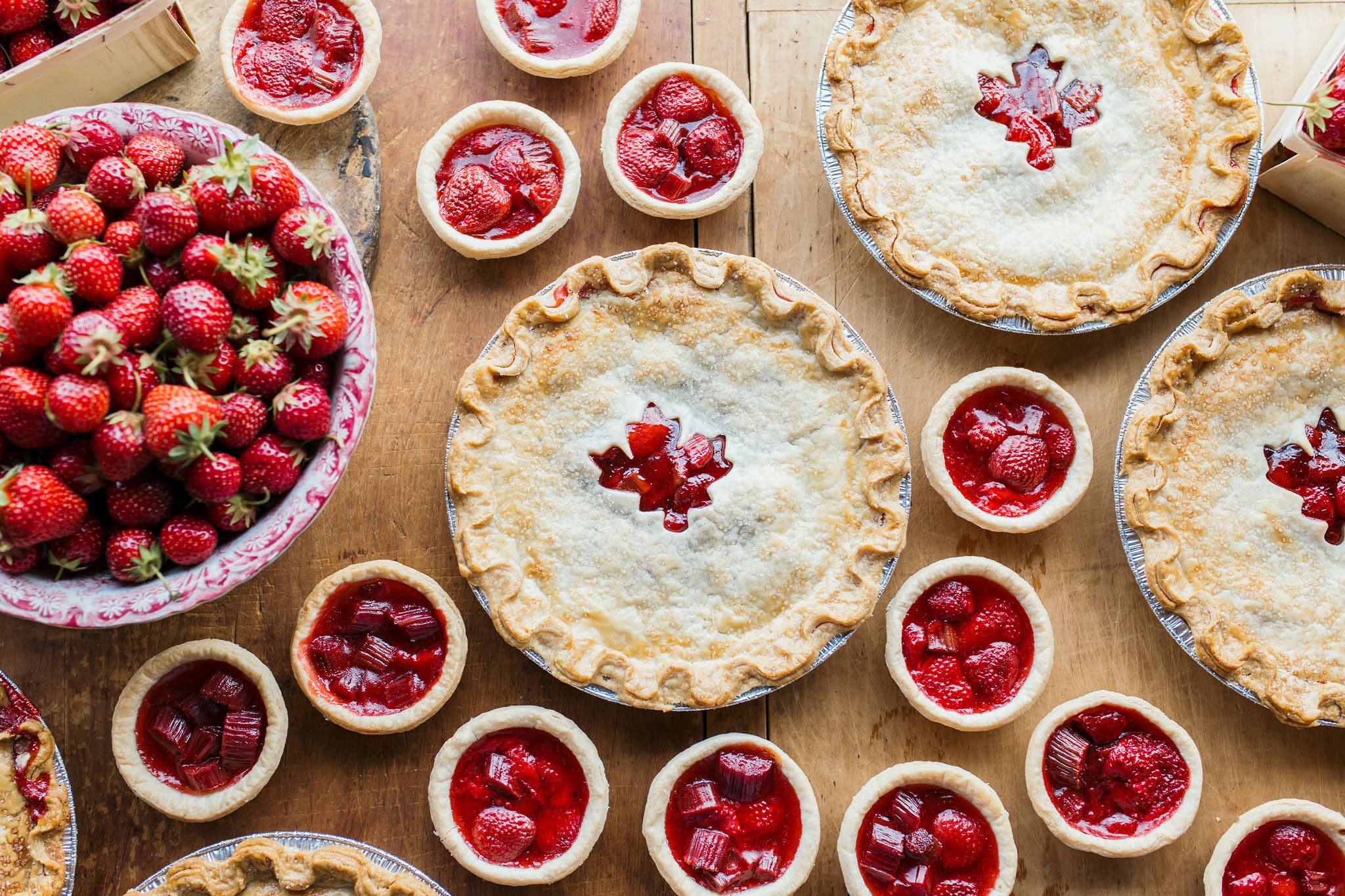 Strawberry pies with maple-leaf cut-outs and rows of strawberry and rhubarb tarts beside a bowl of fresh strawberries