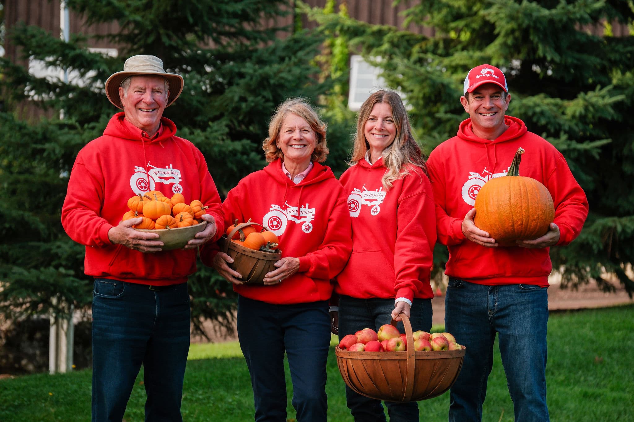 Four smiling members of the farm family in red Springridge hoodies hold bowls of pumpkins and gourds, a basket of apples and a large pumpkin