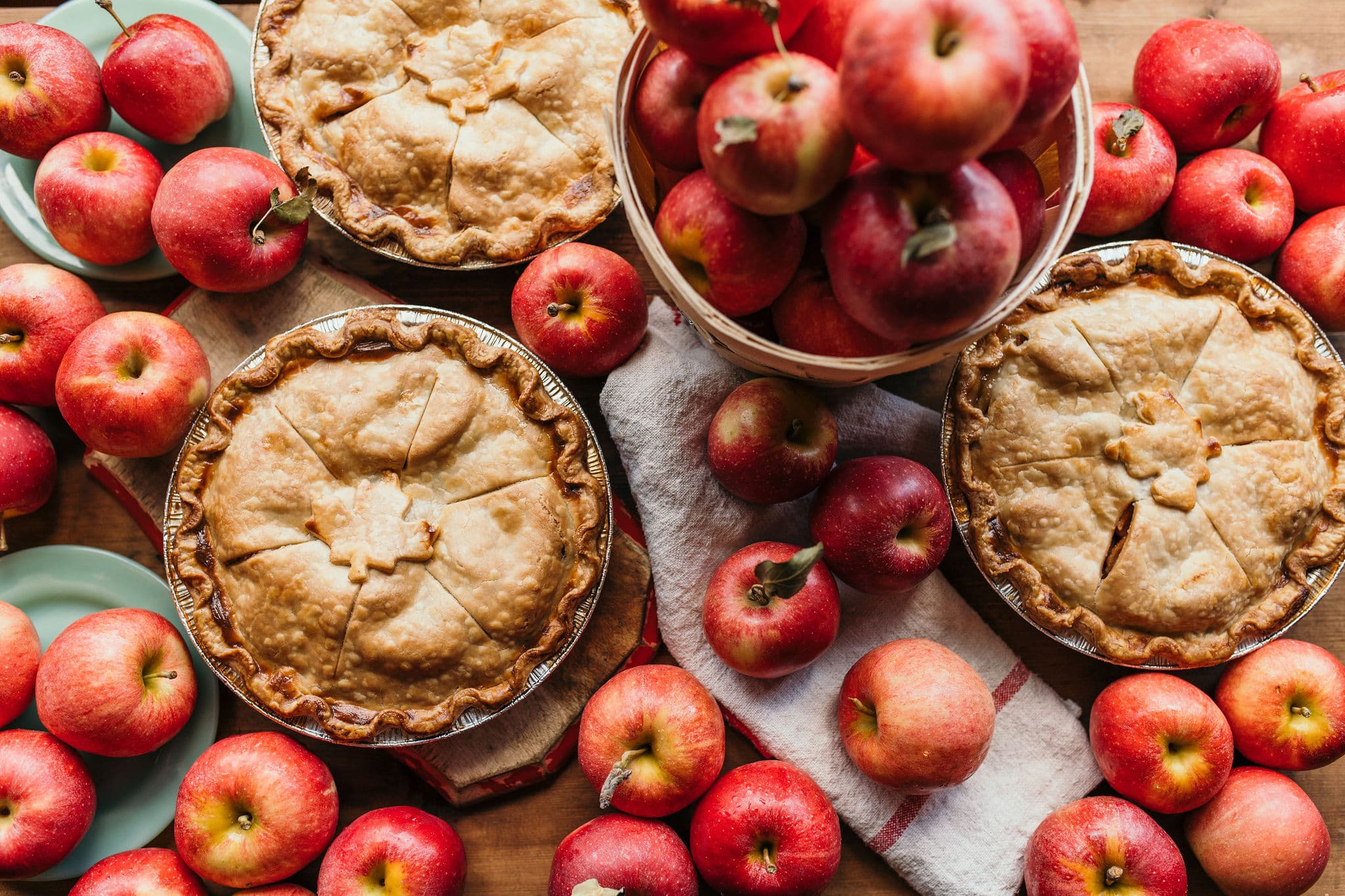 Three golden apple pies on a wooden table among red apples, with a basket of apples and a striped tea towel