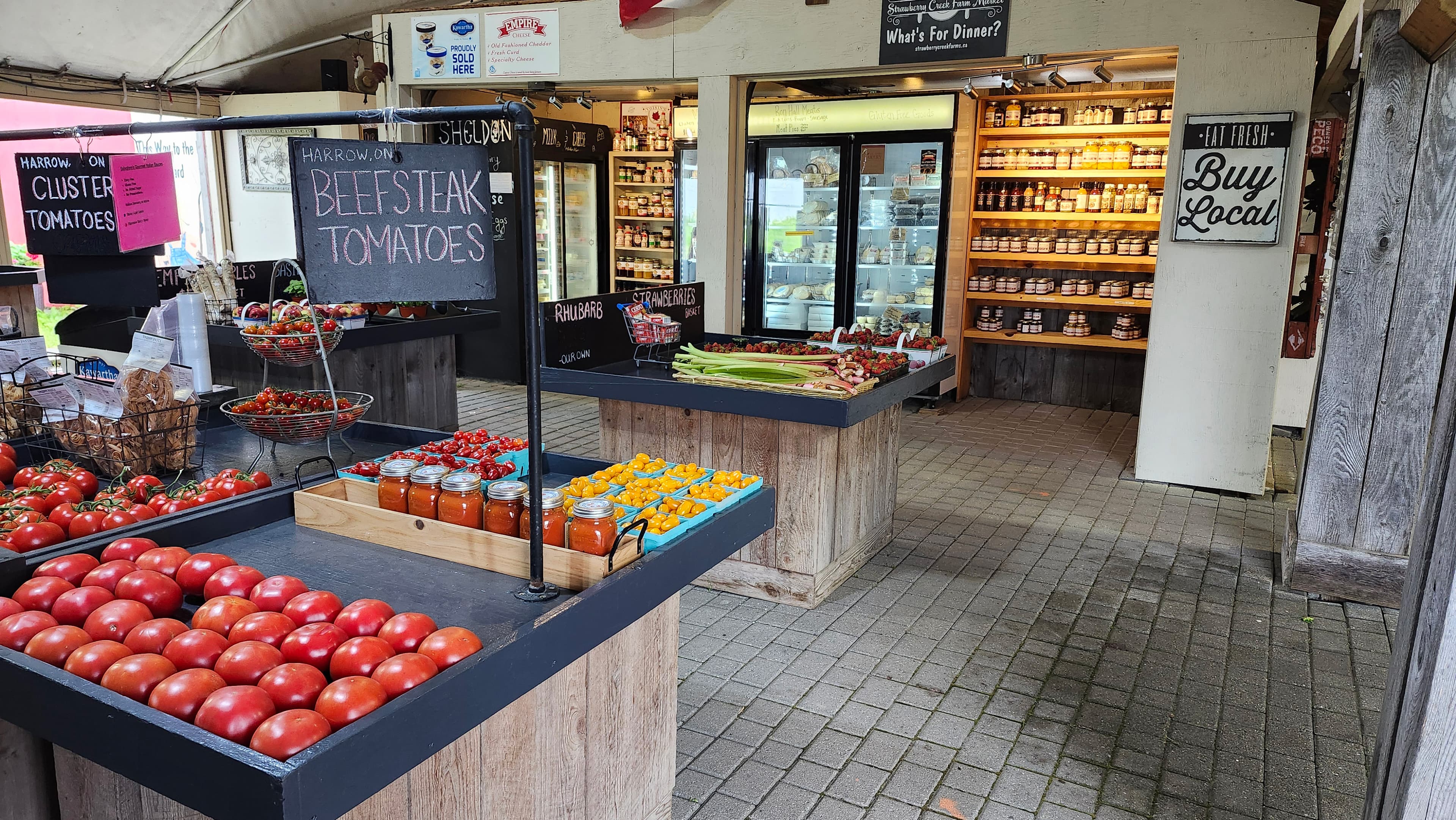 Inside the farm market: tables of tomatoes, rhubarb and quarts of strawberries in front of shelves of preserves and fridges of cheese and baking