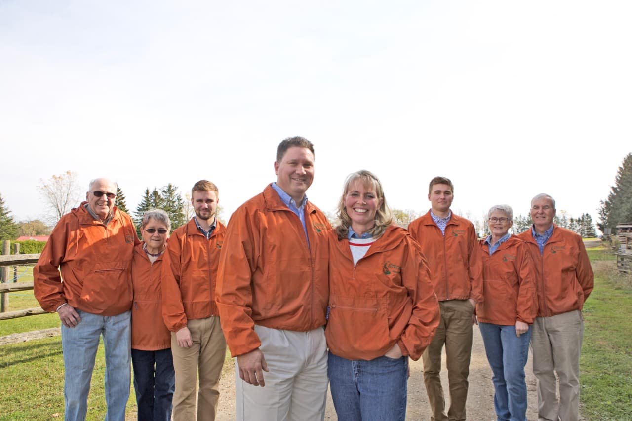 The Strom family, eight adults of three generations in matching orange Strom's jackets, stand smiling on a gravel farm lane beside a split-rail fence