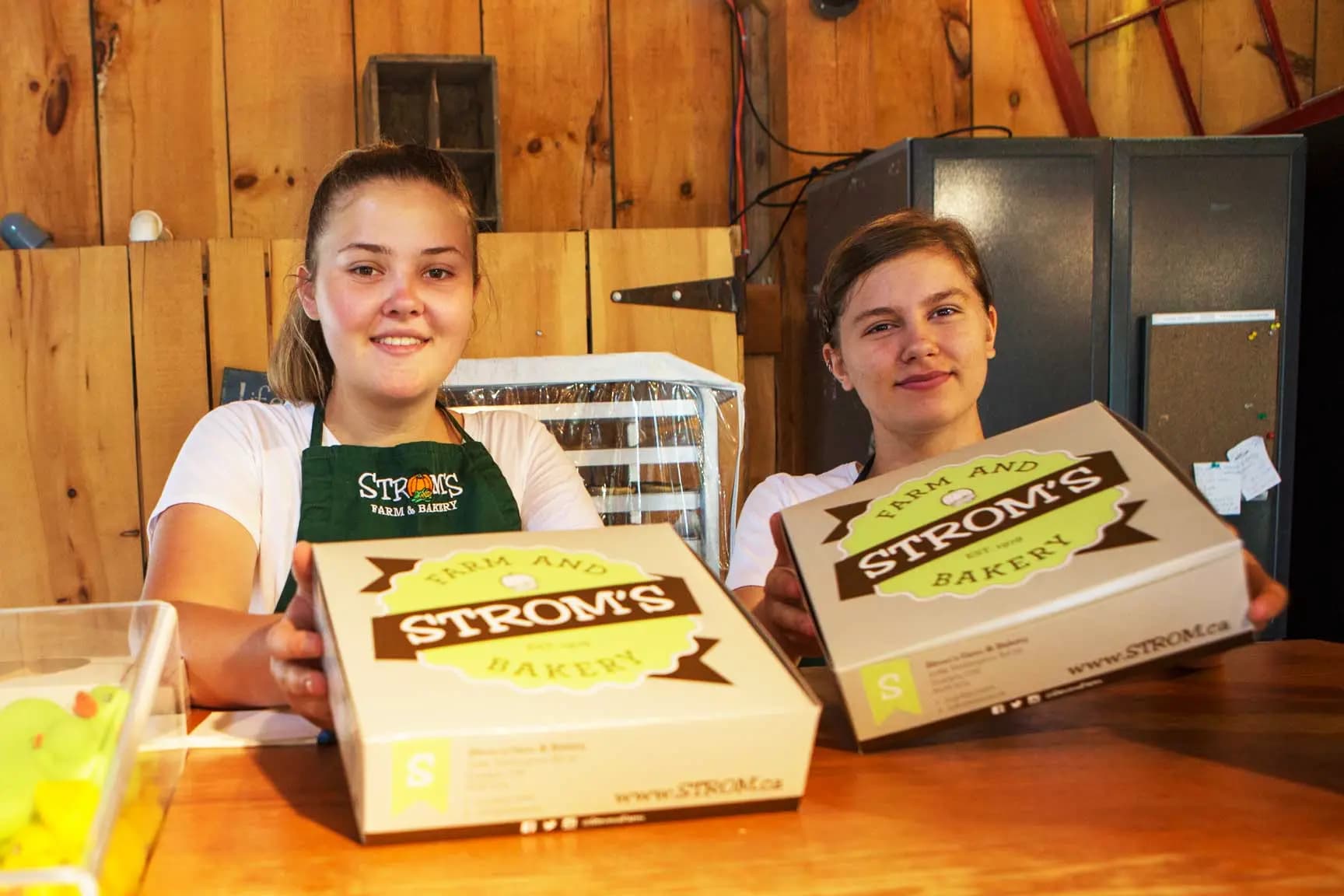 Two bakery staff, one in a green Strom's apron, hold out Strom's Farm and Bakery pie boxes across a wooden counter