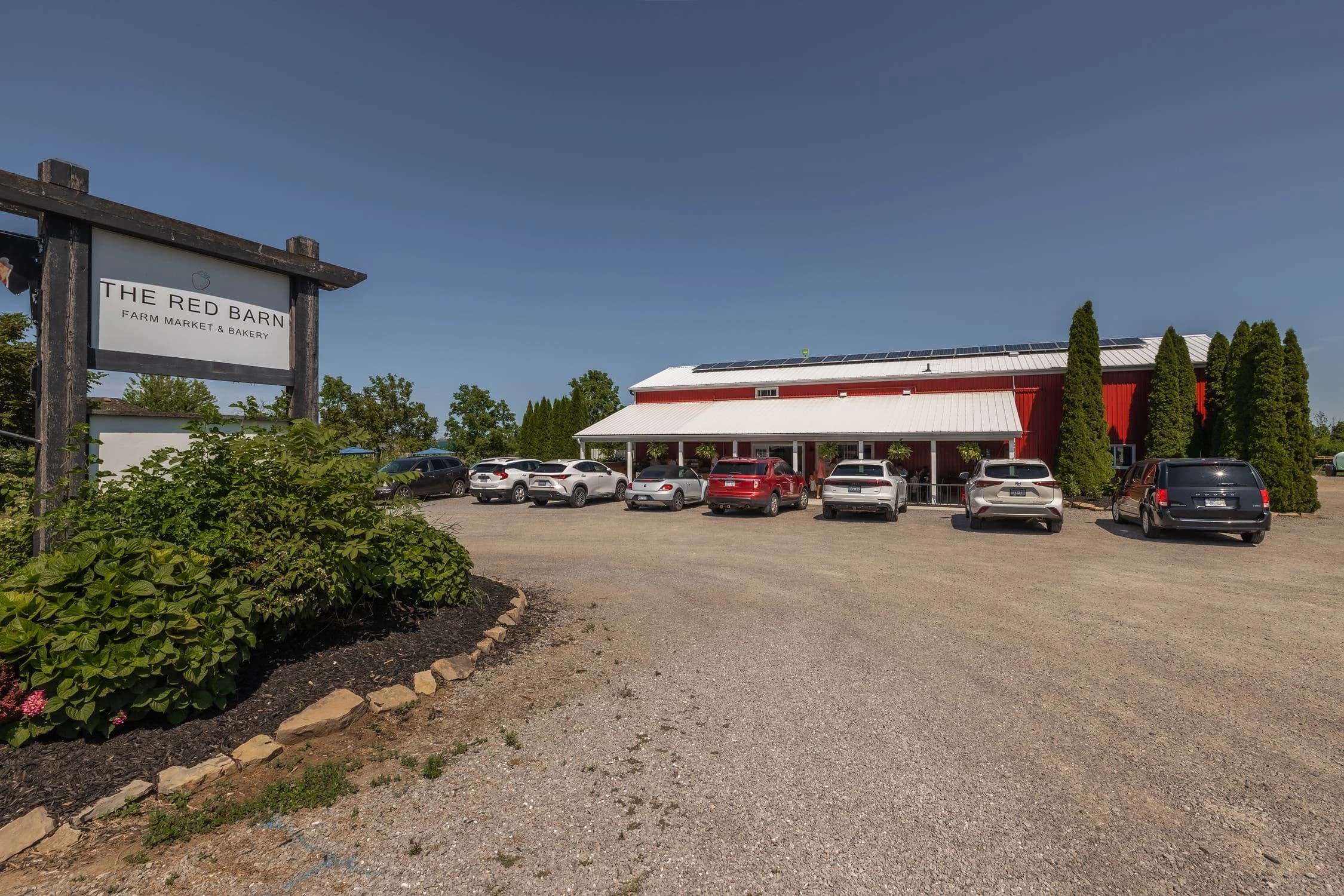The Red Barn Farm Market and Bakery, a long red barn with a white porch roof and solar panels, beside its roadside sign and gravel parking lot