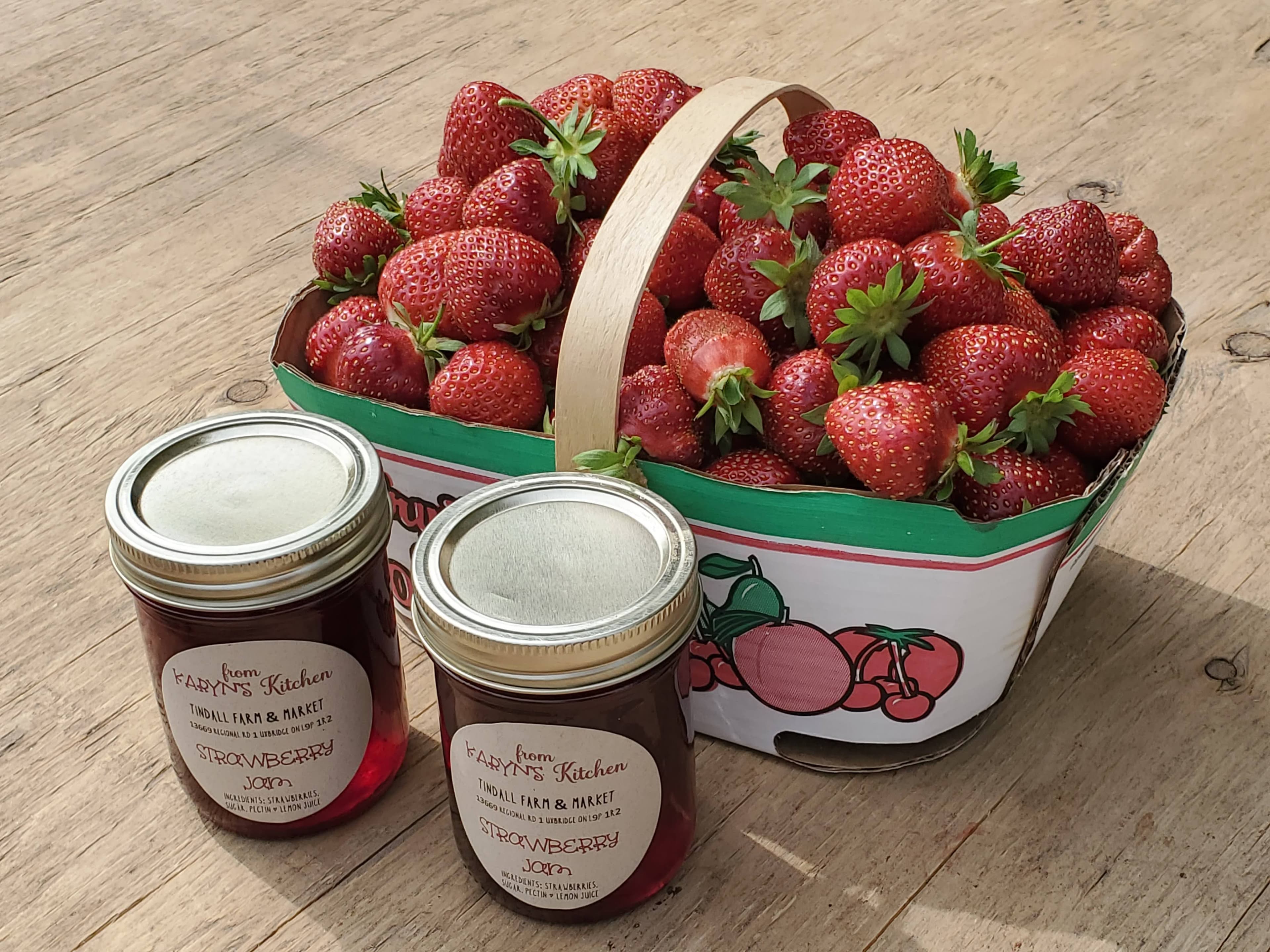 A heaped basket of freshly picked strawberries beside two jars of the farm's own strawberry jam on a sunny wooden table at Tindall Farm & Market