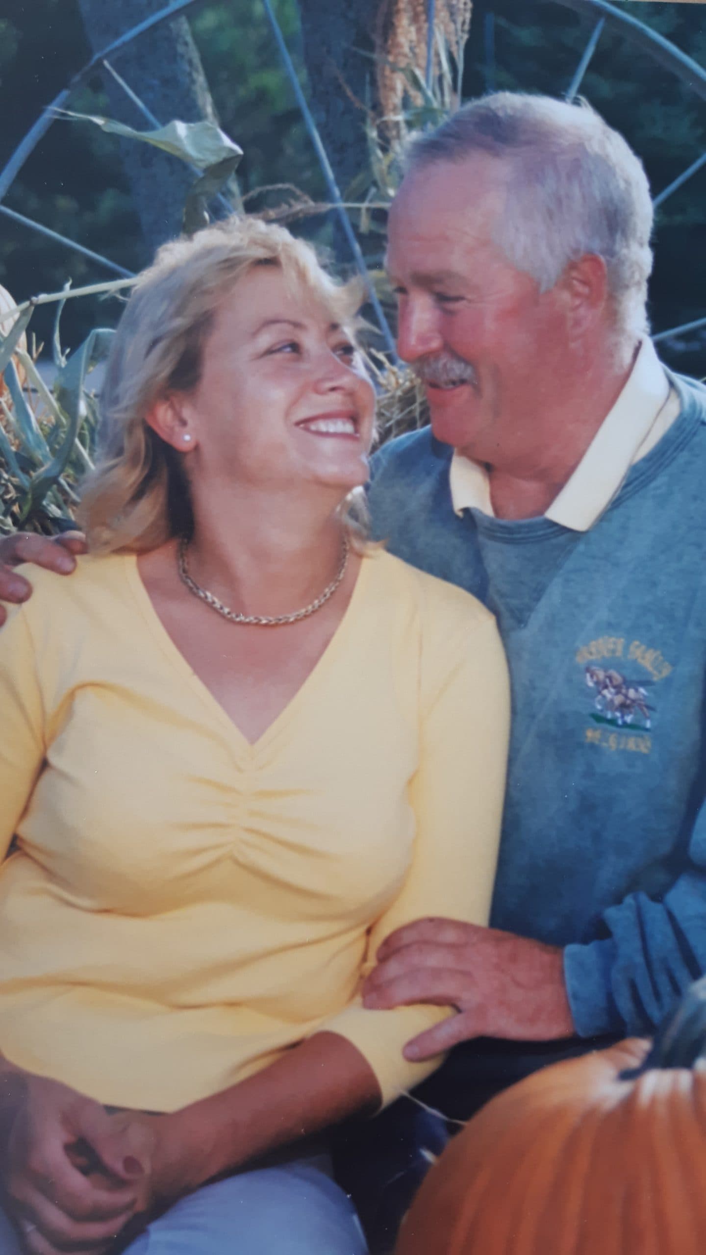 A smiling couple sit together beside a pumpkin and corn stalks in front of an old wagon wheel