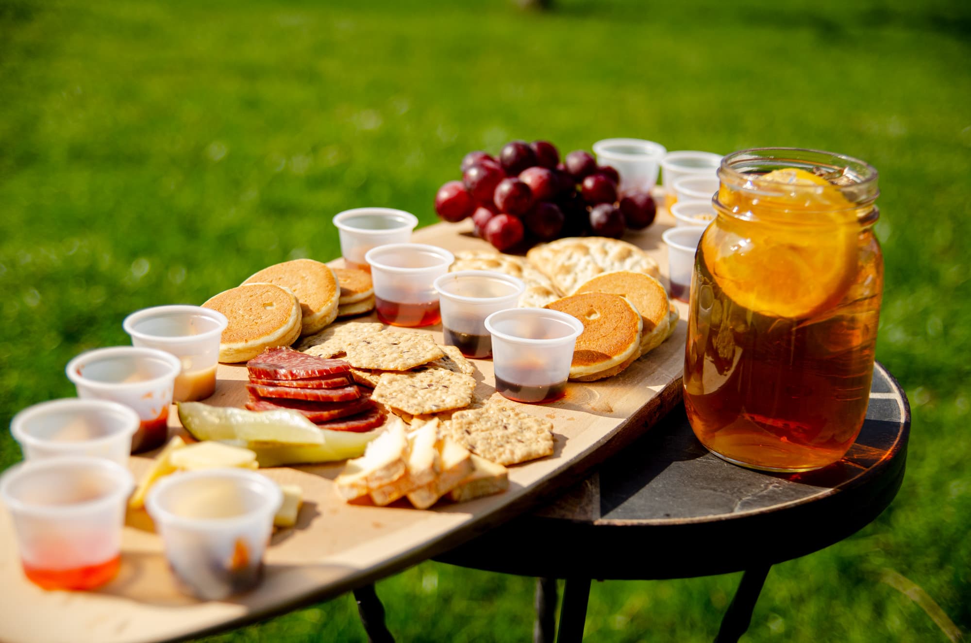 A maple tasting board on a table in the grass: mini pancakes, crackers, summer sausage, cheese, a pickle, red grapes, small cups of maple syrup and a jar of iced tea with an orange slice