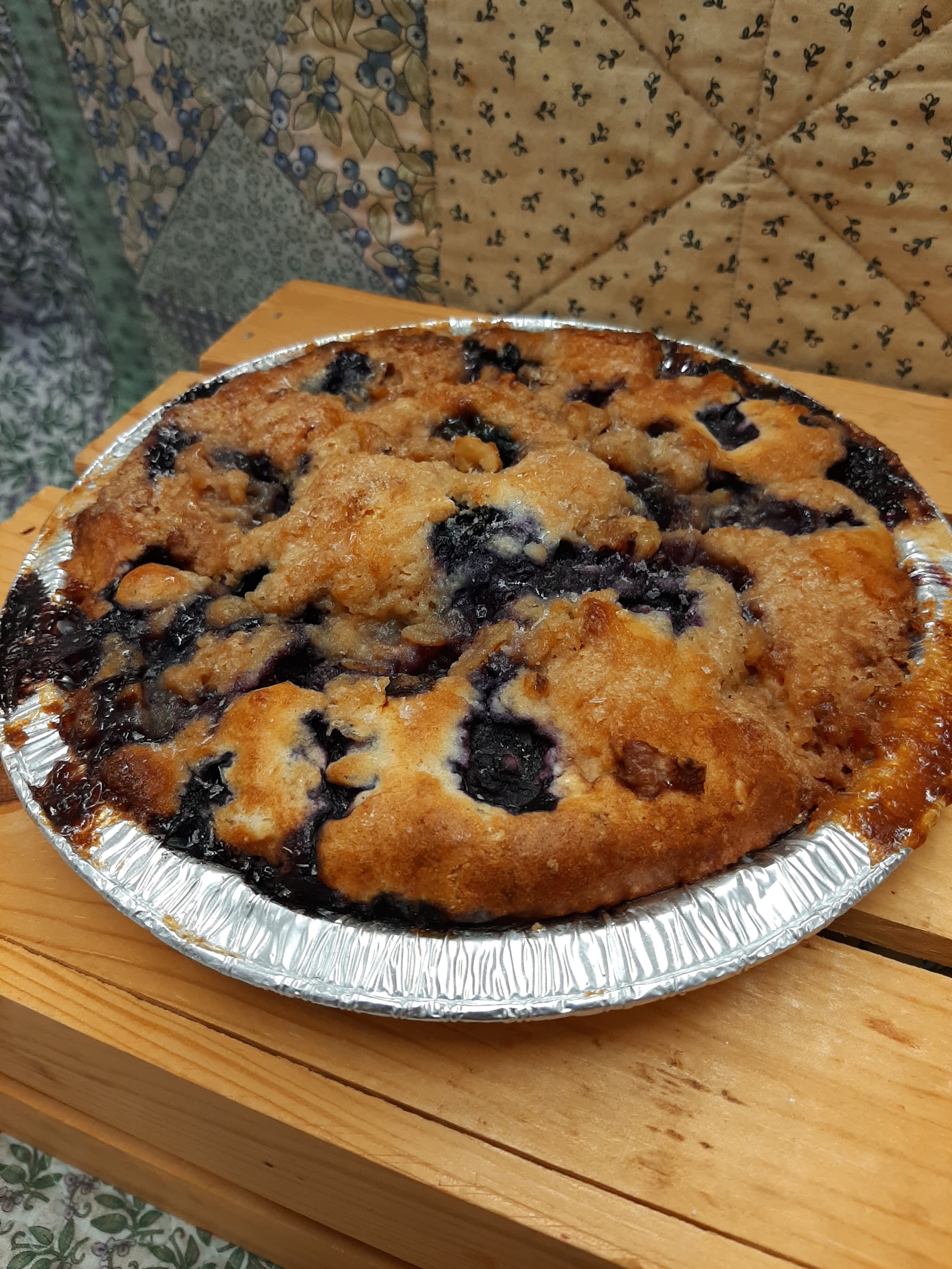 A golden blueberry coffee cake in a round foil pan, set on a wooden crate in front of a patchwork quilt