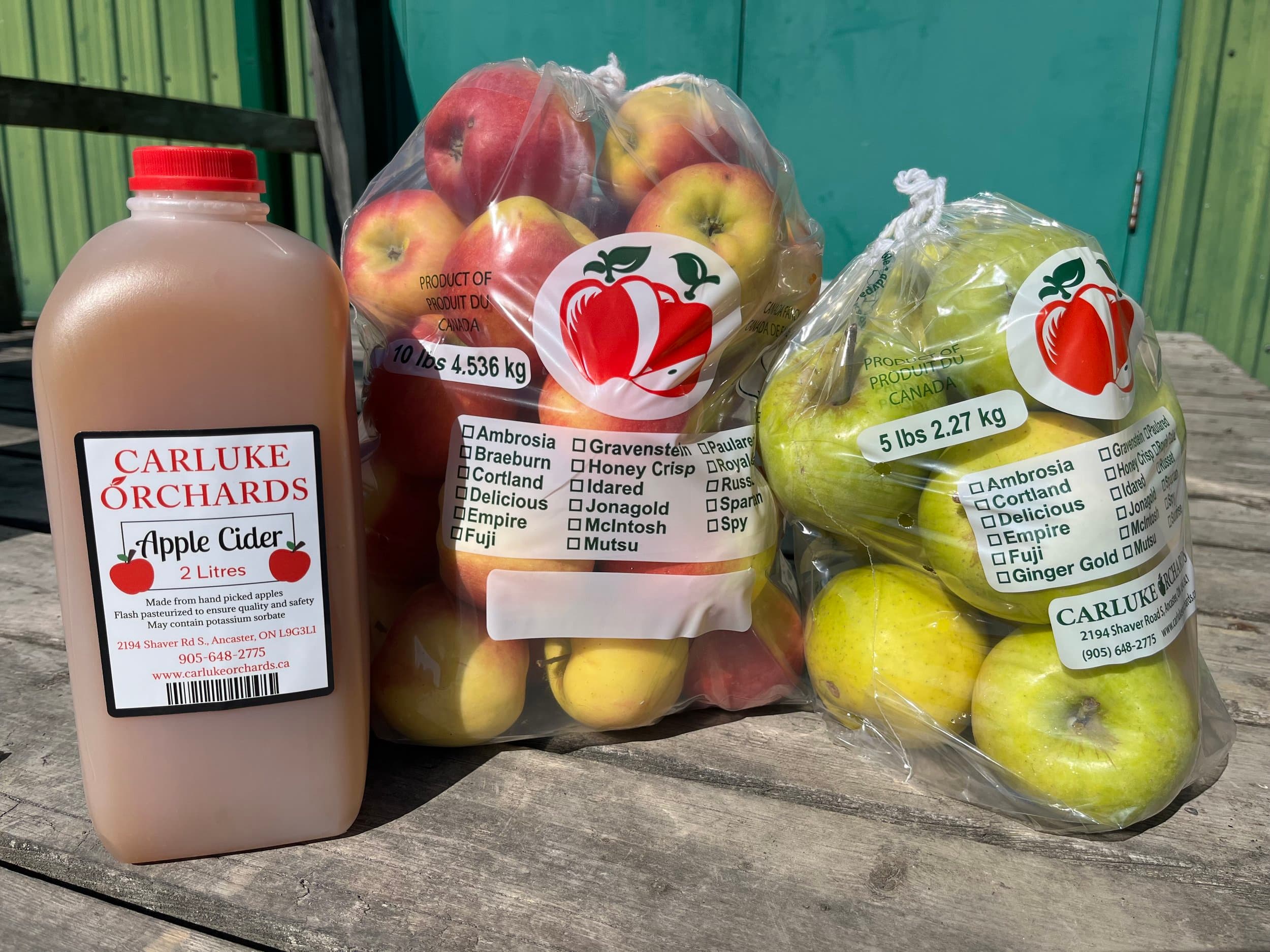 A 2-litre jug of Carluke Orchards apple cider beside a 10 lb bag of red apples and a 5 lb bag of green apples on a weathered picnic table