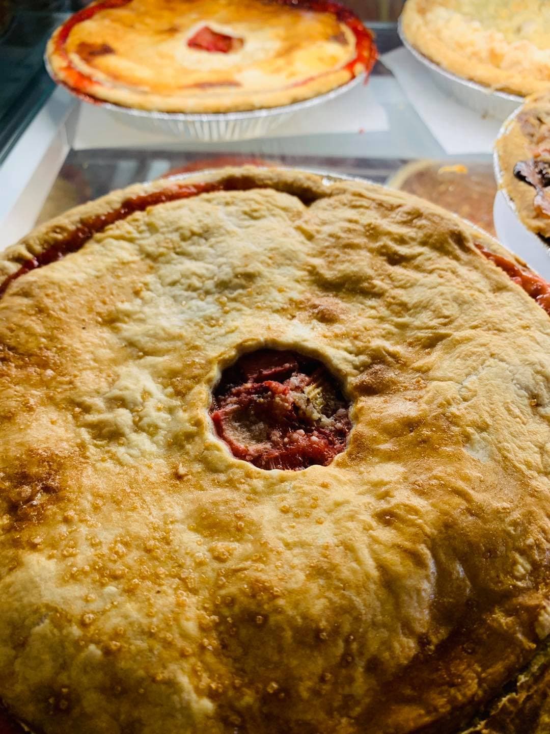 A double-crust fruit pie with a vent cut in the top, with more pies behind it in the bakery's display case