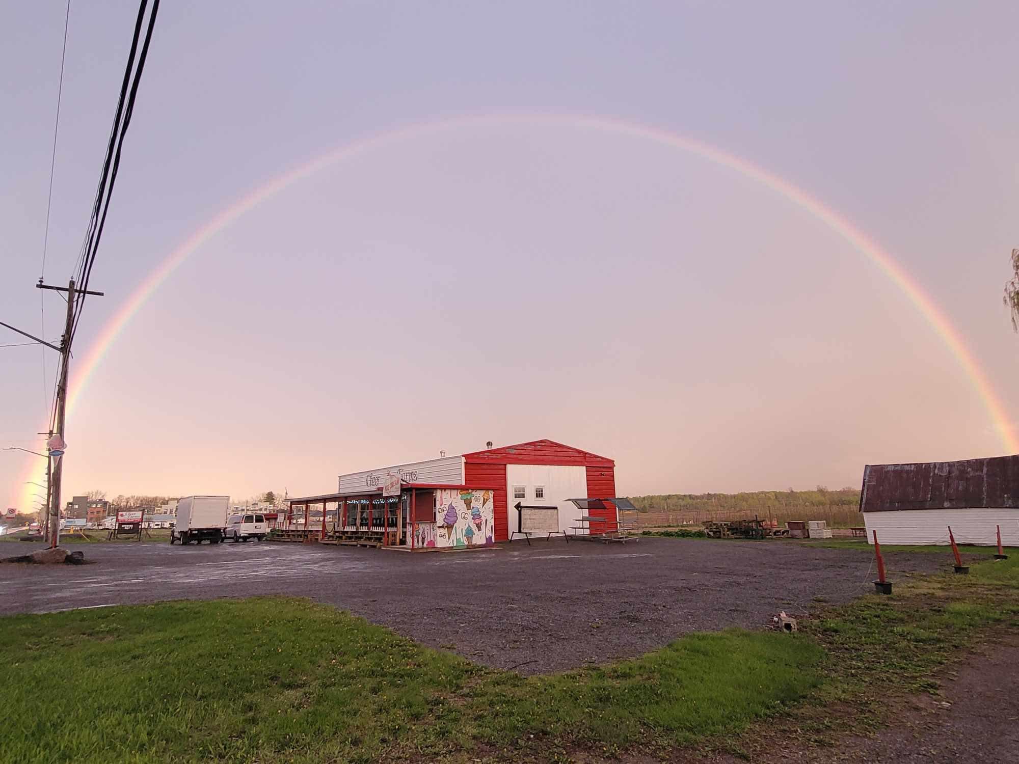 The red and white Cheer Farms market building on Main Street in Brighton, with its painted ice cream mural and covered porch, under a full rainbow