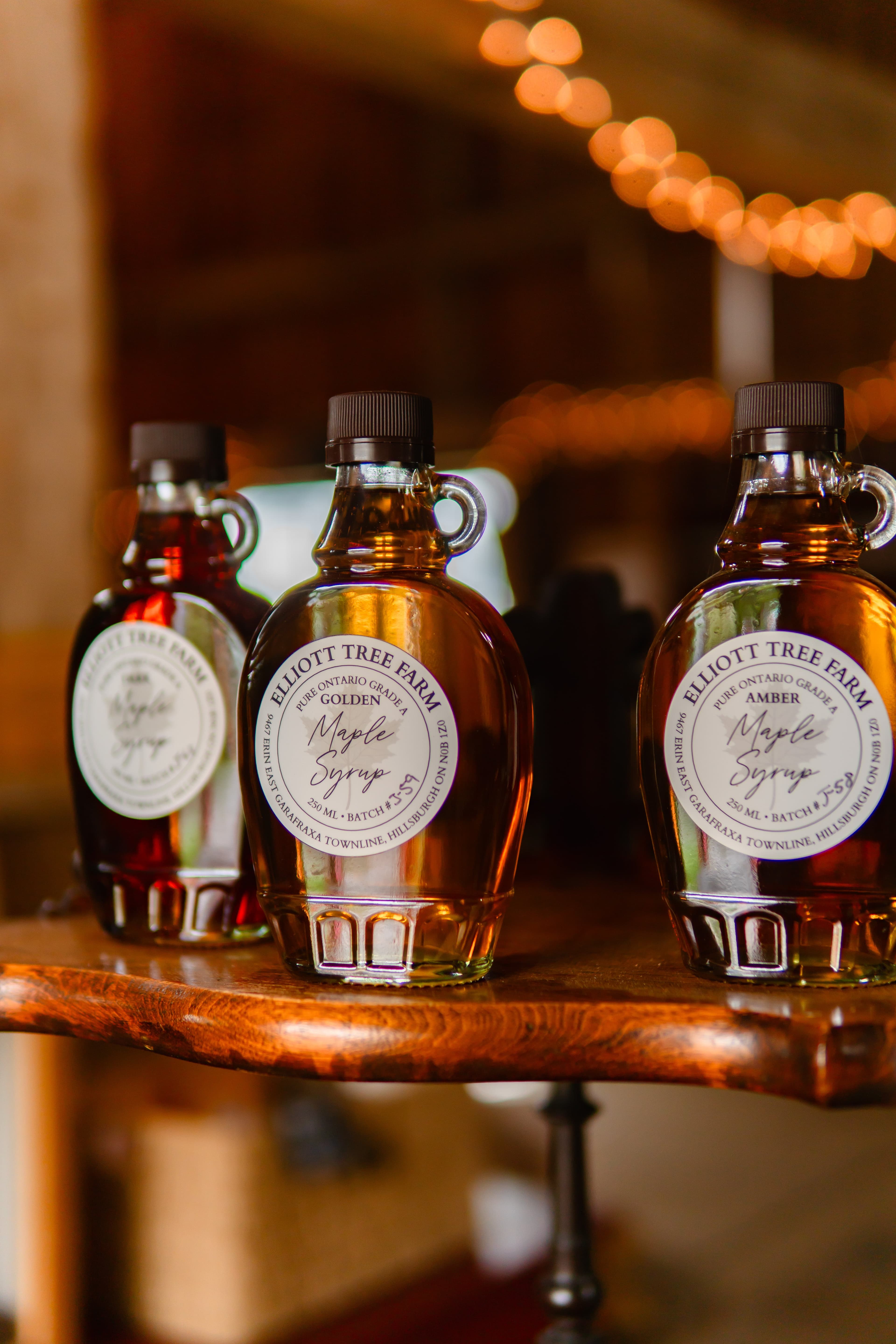 Three bottles of Elliott Tree Farm maple syrup, Golden and Amber labels in front, on a wooden shelf, with string lights glowing in the barn behind