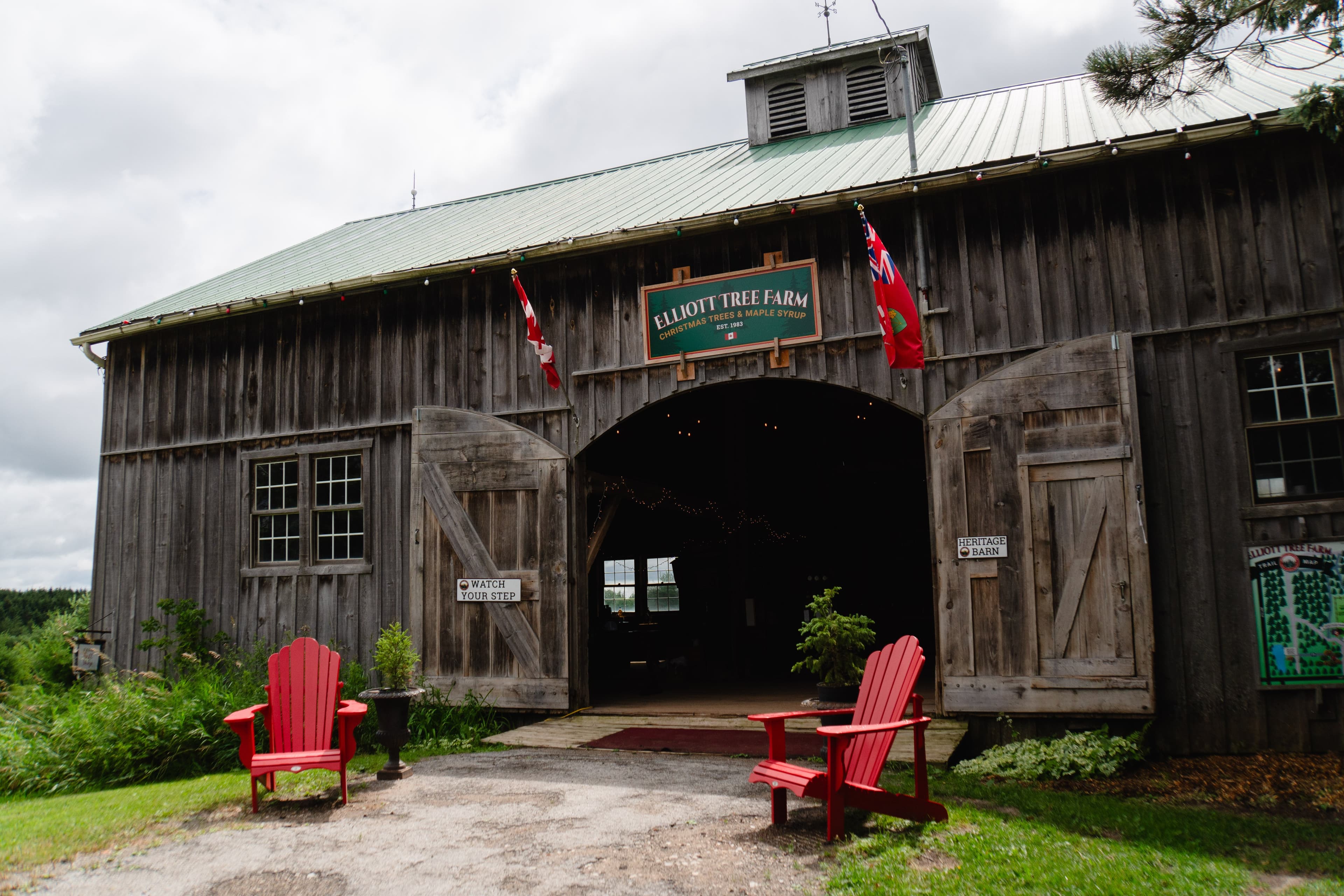 The weathered board-and-batten Heritage Barn with its green Elliott Tree Farm sign over the open arched doors, Canadian and Ontario flags, and two red Muskoka chairs on the lawn